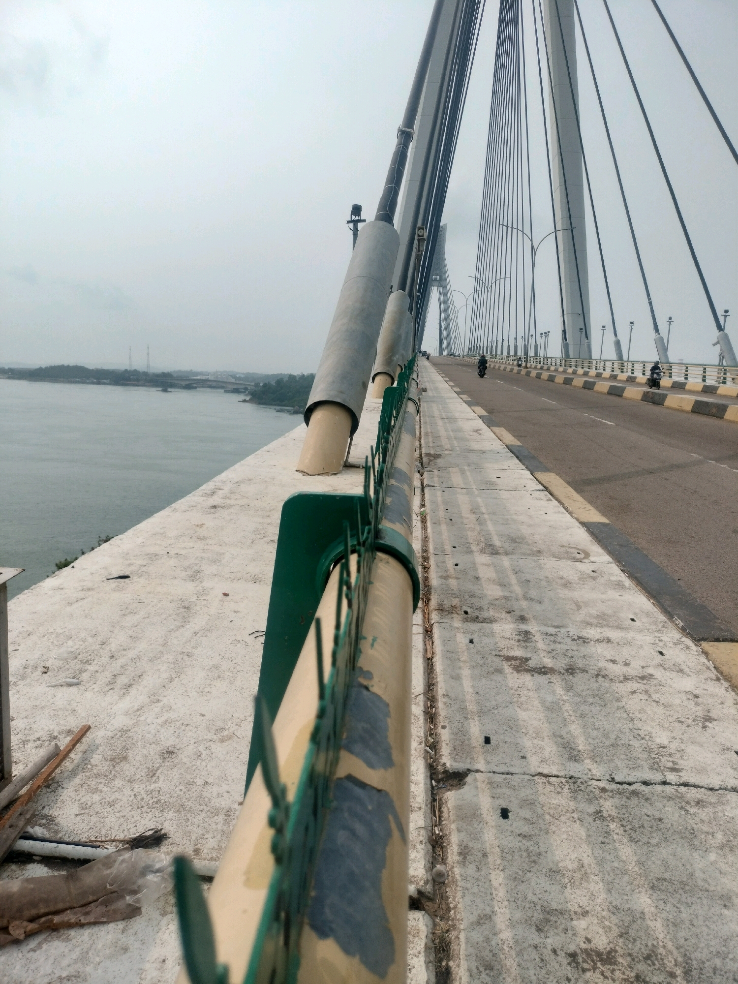 The image shows a perspective view from the pedestrian walkway of the Barelang Bridge in Batam, Indonesia. The bridge is a cable-stayed bridge with a highway that is open to traffic. Two motorcycles are visible on the highway, with riders wearing helmets. The sky is overcast, and the water of the strait is calm. The pedestrian walkway is made of concrete and has a railing that appears to be made of metal and painted green and yellow. There are some construction materials, including wooden planks and plastic sheeting, on the walkway near the foreground. The bridge's large concrete pylons and numerous suspension cables are prominent features in the background.