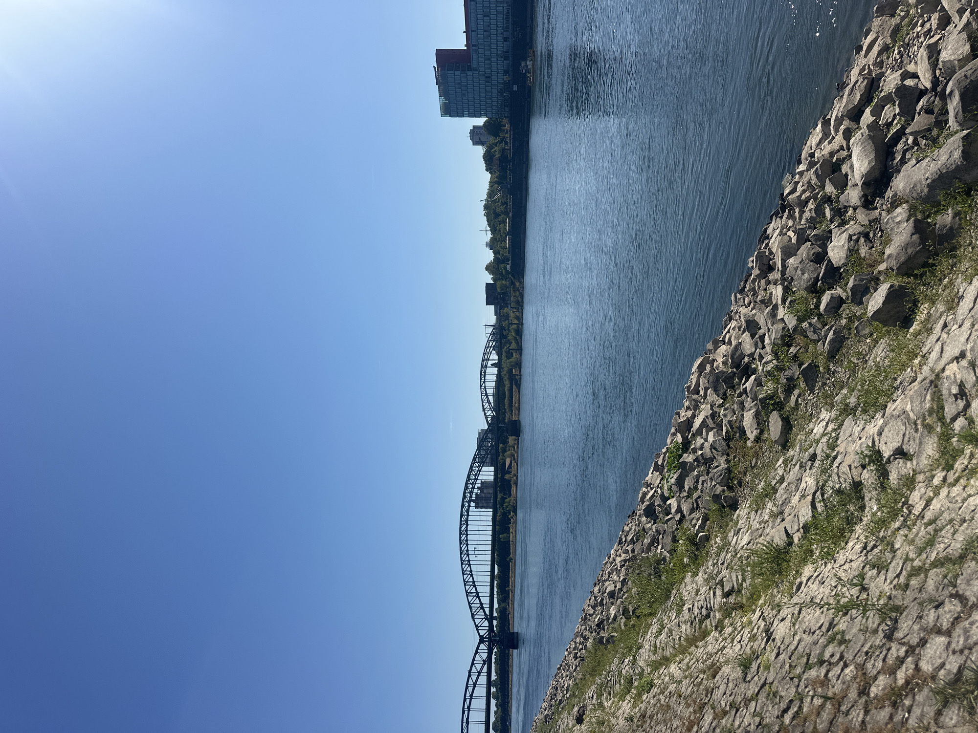 An outdoor, eye-level, wide shot captures a bright, clear day along the Rhine River in Cologne, Germany. The river dominates the right side of the frame, its surface a deep blue, reflecting the clear sky. The left bank of the river is a sloping embankment covered in large, grey rocks and scattered patches of green grass and weeds. Further inland, a multi-arched train bridge spans the river. On the far bank, a tree-lined embankment is visible, punctuated by various buildings. Prominently, a modern, multi-story building with a red section at the top and a glass facade stands on the right side of the far bank. The sky is a vibrant, cloudless blue. No people are visible in the scene.