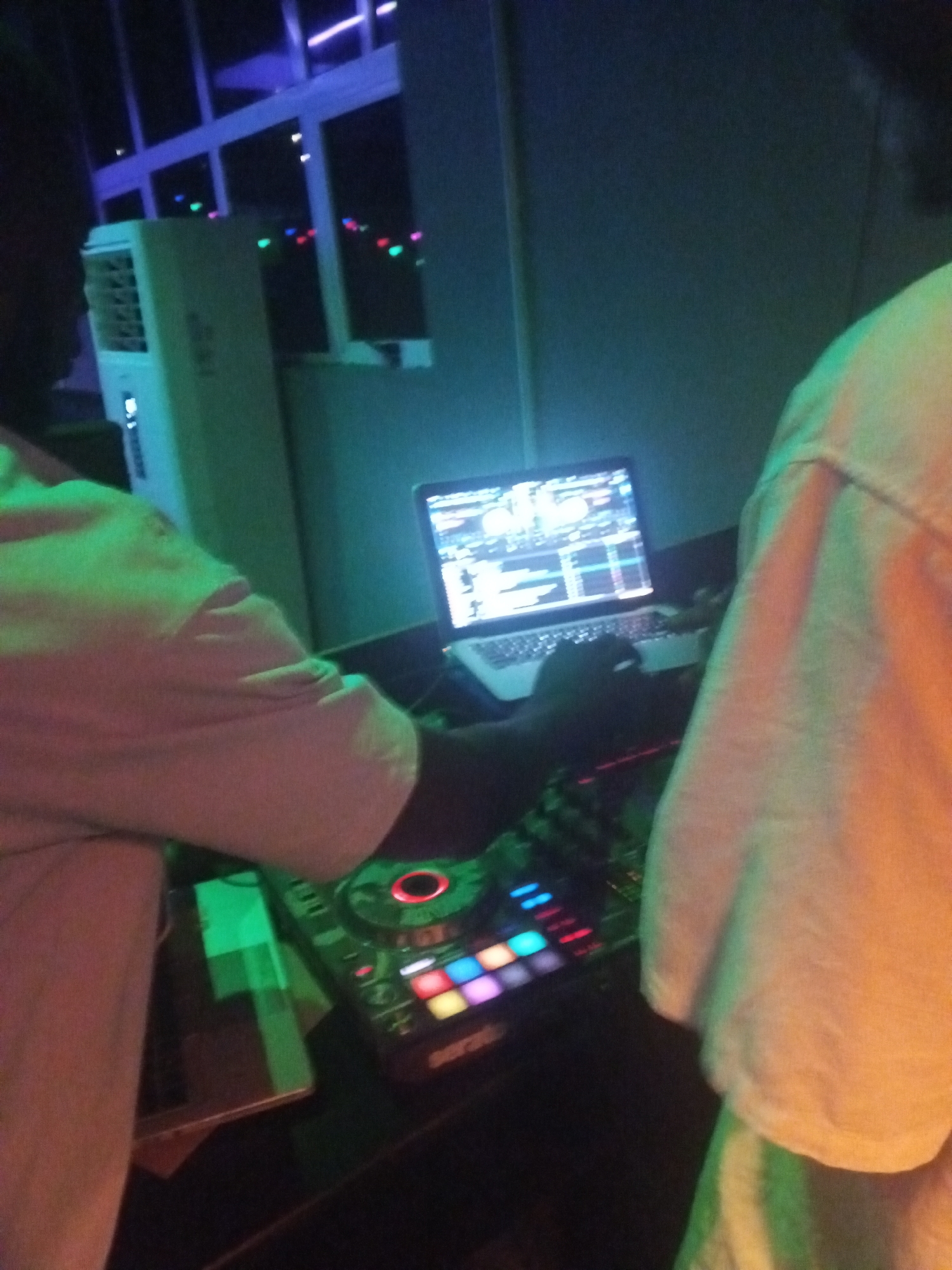 The image depicts a DJ setup in a dimly lit indoor environment, possibly a club or party venue in Unknown, Unknown. A laptop with DJ software is visible on a table, adjacent to a DJ controller with illuminated buttons and knobs. A person's arm is visible operating the controller, while another person is seen partially at the right. An air conditioning unit and a window with colorful lights outside are in the background.
