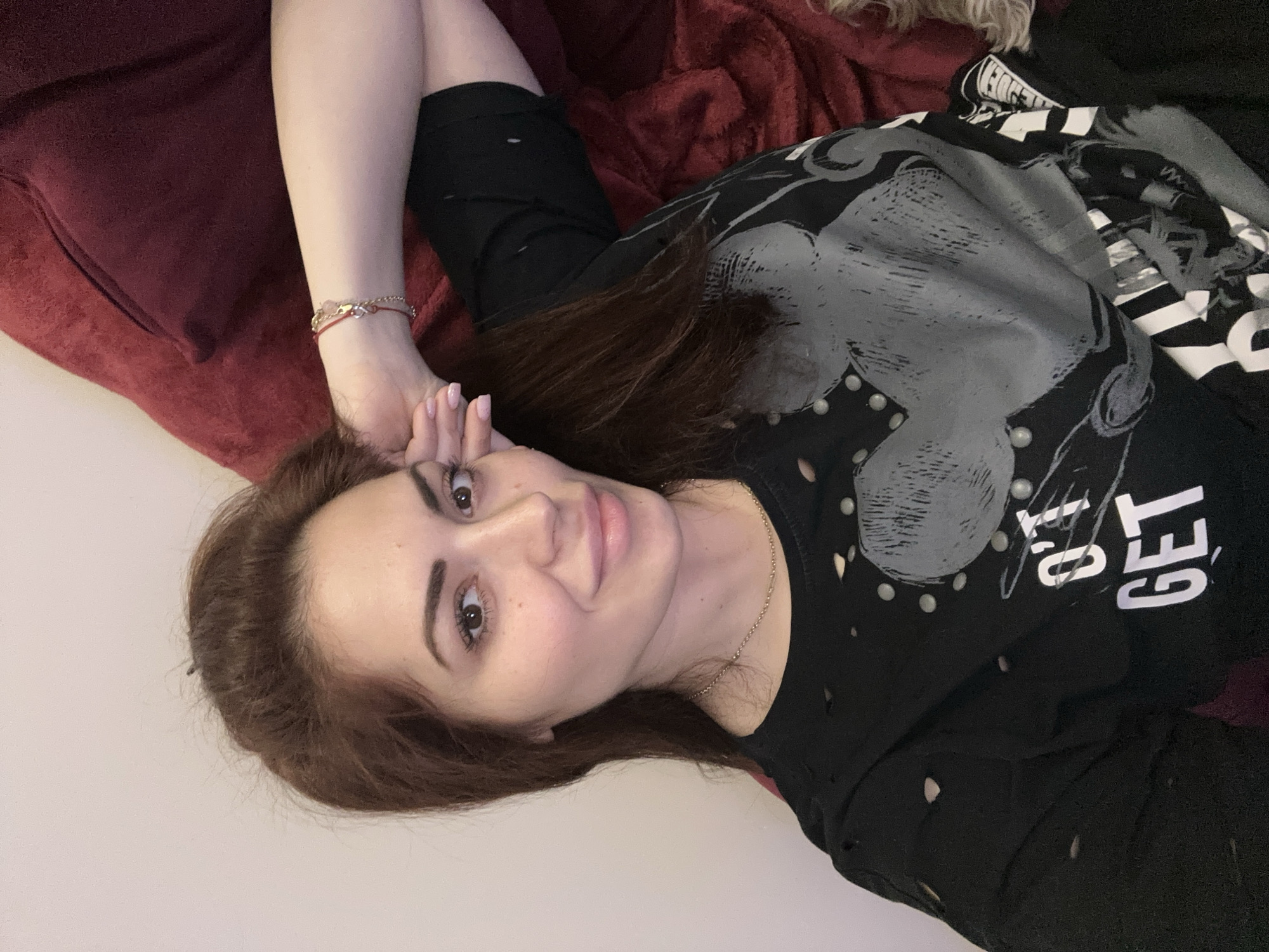 A woman is depicted lying down indoors, looking directly at the camera with a soft, inviting smile. She has long, dark brown hair that cascades around her, fair skin, and prominent dark eyes. Her left arm is bent, with her hand gently resting near her head.
She is wearing a black long-sleeved shirt with a distressed aesthetic, characterized by numerous small holes. The shirt features a large grey graphic on the chest, which appears to be a stylized head or design, adorned with small white, circular details resembling studs or pearls. Partially visible white text, reading "OT GET," is printed on the shirt over the graphic. She accessorizes with a delicate gold-colored chain necklace and a small red and gold-colored bracelet on her left wrist.
The woman is resting on a textured, reddish-brown fabric, likely a blanket or bedding, suggesting a relaxed indoor setting. A plain, light-colored surface or wall is visible behind her. The overall impression is one of calm and casual comfort. The image was taken in Sofia, Bulgaria.