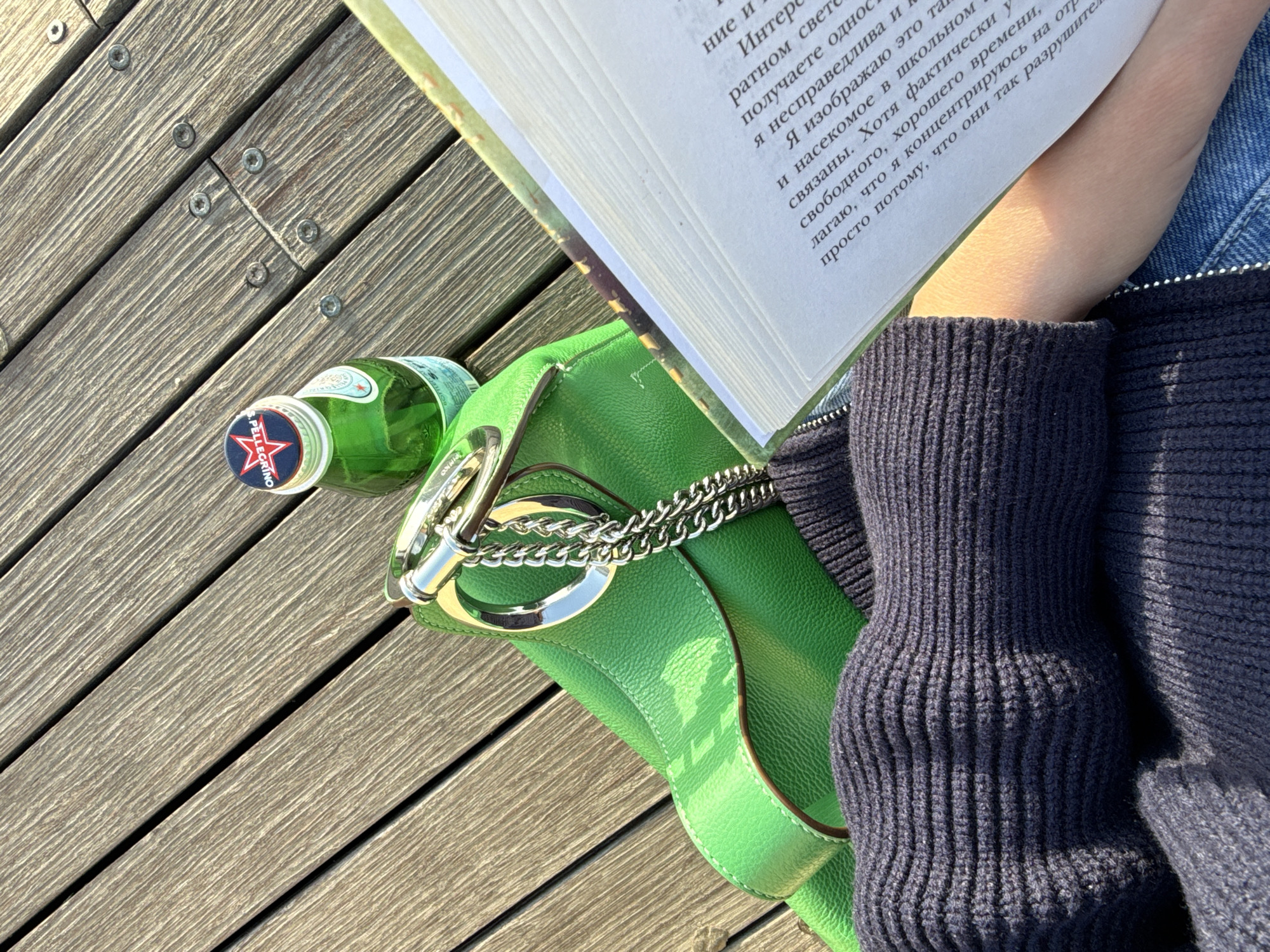 This image depicts a person reading a book outdoors, accompanied by a green purse and a bottle of San Pellegrino water. The scene is set on a wooden deck, likely a patio or boardwalk. The person is wearing a dark blue knit sweater and denim jacket. The book's open pages display text in a language other than English. The purse features a silver chain and hardware. The San Pellegrino bottle is positioned on its side, with the logo visible. The location appears to be outside, with natural light illuminating the scene. Given the context, it's possible that the location is Tel-Aviv, Israel, as the prompt suggests, although without further information this location cannot be confirmed.
