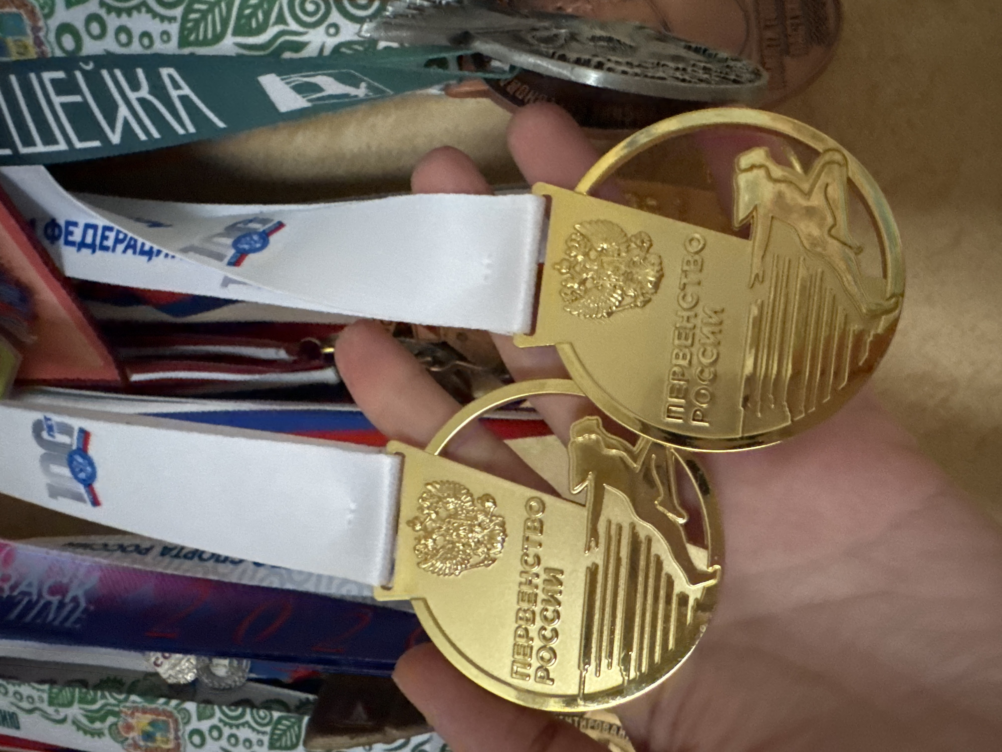 In this close-up image, a pair of gold medals hangs from white ribbons, held in a person's hand. The medals are circular, with embossed designs. The upper part of each medal features a double-headed eagle crest. Below the crest, the Russian text "ПЕРВЕНСТВО РОССИИ" (Championship of Russia) is inscribed. A stylized silhouette of a runner is depicted in motion, with horizontal lines suggesting speed. The background is a jumble of other medals and ribbons, displaying various colors and patterns. One ribbon on the left has a green pattern with white circles, while another on the right is dark green with white lettering. The overall scene suggests a sporting event or achievement. The provided coordinates, while specific to Sinyavino, Russia, do not offer direct visual confirmation from the image alone; their relevance is implied through the context of the medals' inscriptions.
