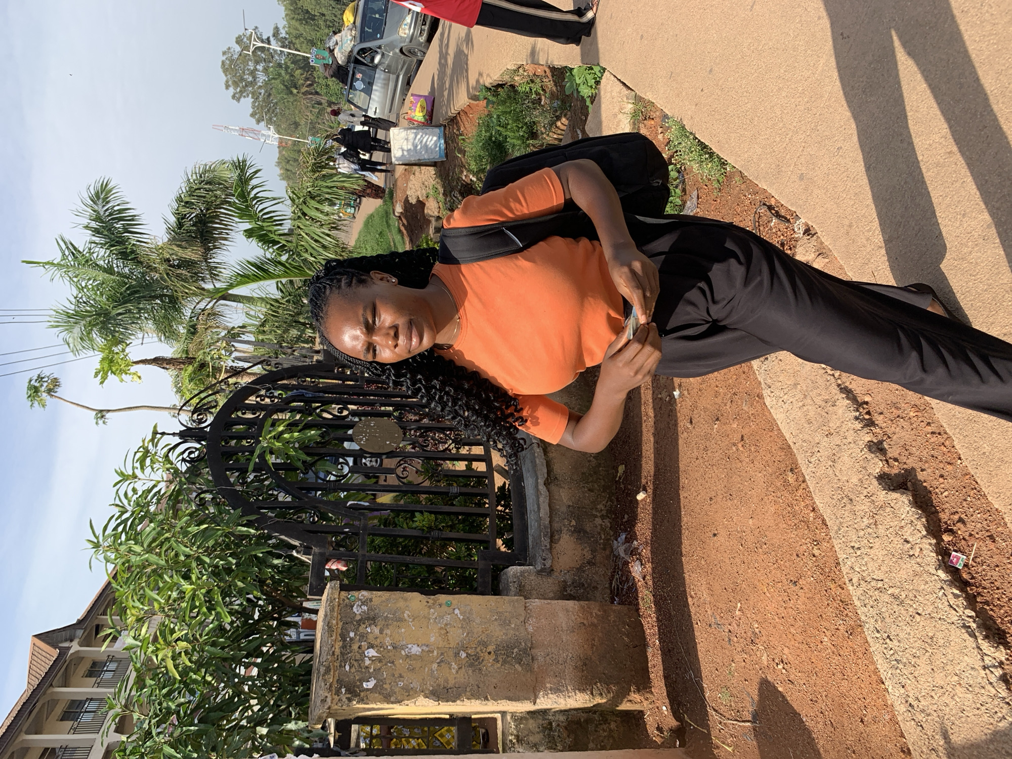 A young African woman with dark, curly hair styled in braids walks on a concrete sidewalk in Gura Top, Nigeria. She is wearing an orange t-shirt and black pants, and a black backpack is slung over her shoulders. In her right hand, she holds what appears to be some paper money. The background features a building with balconies, a large ornate metal gate, lush green trees, and palm trees. Beyond that, a street scene unfolds with several people walking, a silver car loaded with luggage on its roof, and a telecommunications tower visible in the distance. The ground in front of the sidewalk is a reddish-brown dirt. The lighting suggests it is daytime, with shadows indicating a low sun angle.