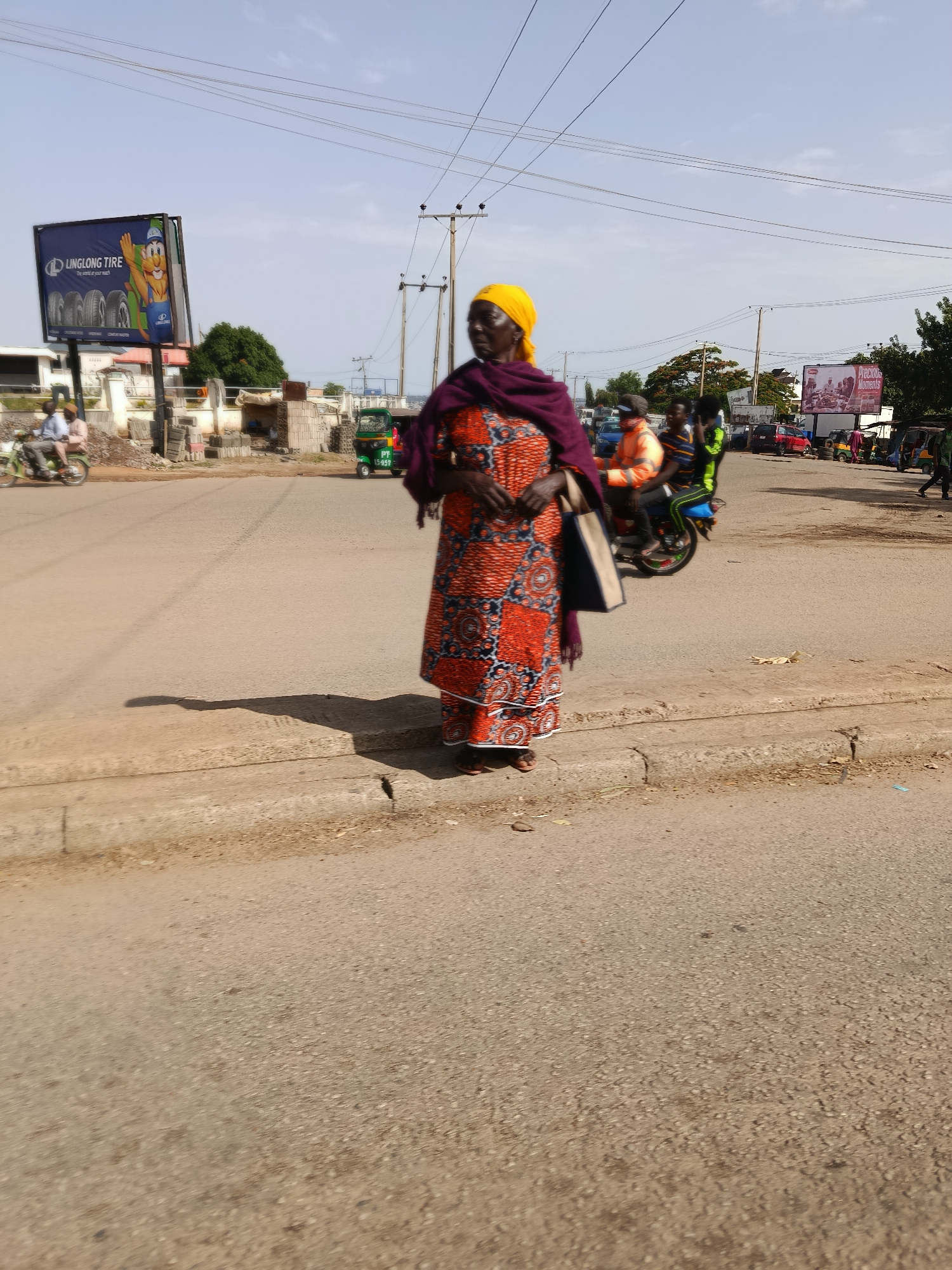 A woman in a vibrant orange and blue patterned dress and a yellow headscarf stands on a street in Jos, Nigeria. She is carrying a dark bag over her right shoulder. The street is paved, with a curb and gutter visible in the foreground. In the background, there are several vehicles, including a green three-wheeled auto-rickshaw with a license plate reading "PT 557 C", motorcycles, and cars. A large billboard advertising "LINGLONG TIRE" is visible on the left side of the image, featuring cartoon characters and tires. Another billboard with the text "Precious Moments" is visible on the right. Power lines crisscross the sky, and utility poles are present along the street. The overall scene suggests a busy urban environment in Jos, Nigeria, with everyday street life and commercial advertising.