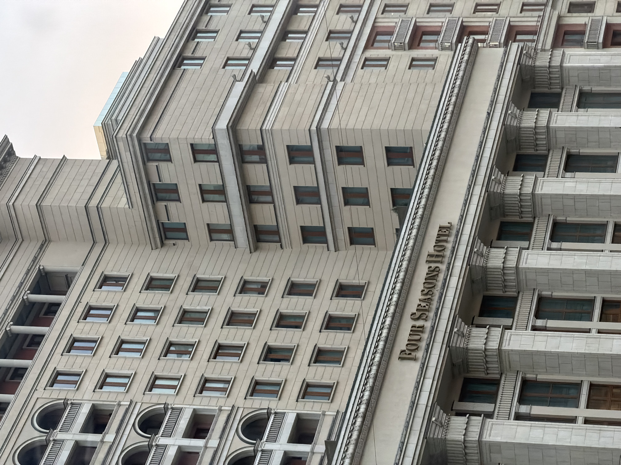 The image depicts the Four Seasons Hotel in Moscow, Russia, from a low-angle perspective. The building's facade is characterized by a repetitive pattern of windows and protruding architectural features, including stepped setbacks and decorative elements. The hotel's name, "FOUR SEASONS HOTEL," is vertically displayed on a prominent section of the building. The sky above is overcast.