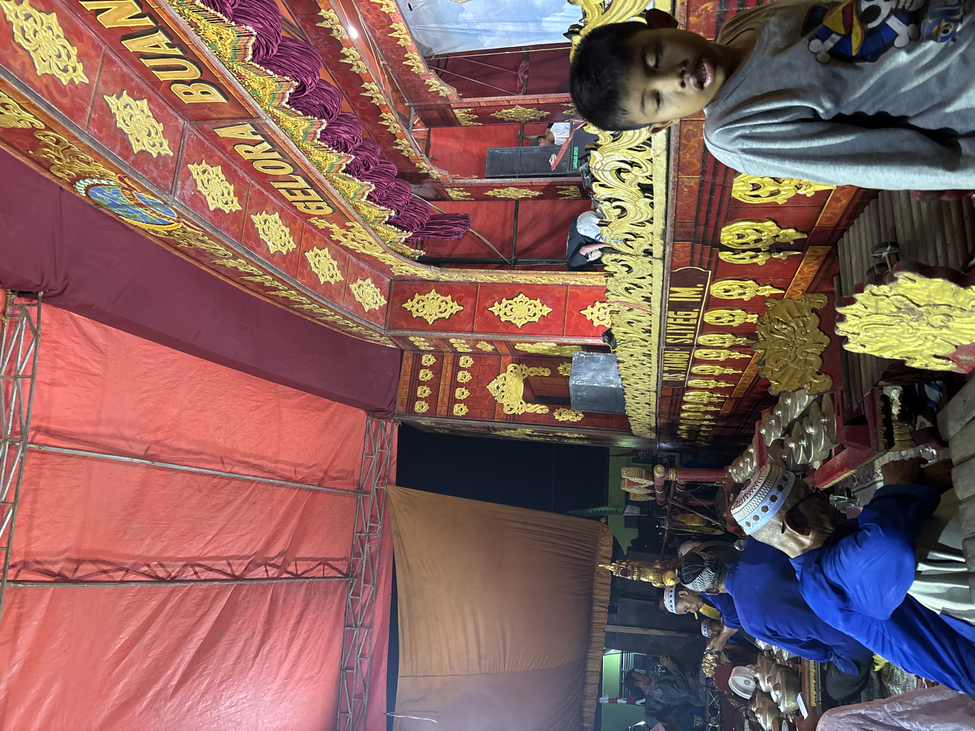 An outdoor event is taking place under a red tent, possibly a performance or gathering, in Panongan, Indonesia. In the foreground, a young boy with dark hair looks to his right with his mouth open. He is wearing a gray long-sleeved shirt with a graphic design on the front. Behind him, a stage is decorated with red and gold ornamentation, with a sign that reads "GELORA BUAN" and "DS. TAMBI SLIYEG, IM.". A group of men in blue shirts and white kufi hats are seated, appearing to play gamelan instruments. One man in the foreground, also in a blue shirt and kufi hat, is looking down. The background shows more of the stage setup with speakers and people working. The environment appears to be illuminated by artificial lights.