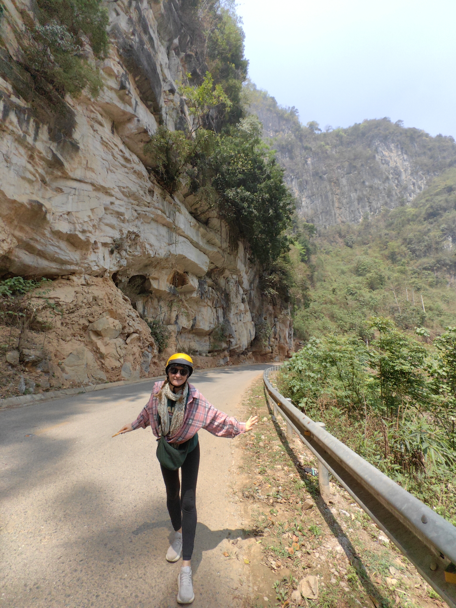 A woman wearing a yellow helmet, sunglasses, a plaid shirt, and black leggings walks along a paved road in Xã Xuân Trường, Vietnam. The road curves around a large, layered rock formation on the left side, which has vegetation growing on it. A metal guardrail runs along the right side of the road, separating it from a lush, green area. Behind the woman, a tall, rocky mountain rises, partially covered in trees and foliage. The sky is light blue, suggesting daylight. The woman has her arms extended to the sides and is facing forward. She wears a scarf and a green hip bag. The road surface appears cracked and uneven. The scene conveys a sense of adventure and exploration in a natural environment.