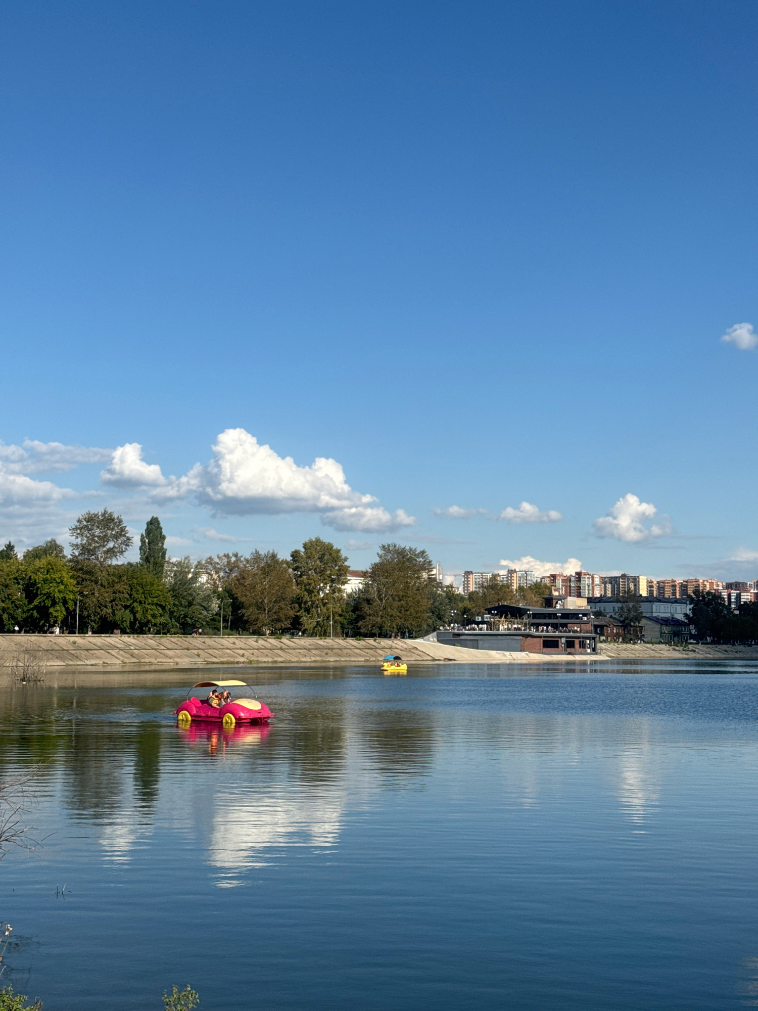 The image depicts a bright, sunny day with a clear blue sky and scattered white clouds, reflected in the calm waters of a lake. In the foreground, a pink pedal boat shaped like a car, with yellow wheels, carries two individuals. Further out on the water, a smaller yellow pedal boat is visible. The lake's edge is lined with a concrete embankment, beyond which a line of trees forms a green buffer. In the mid-ground, a modern building with a dark roof, possibly a restaurant or entertainment venue, is situated on the shore, with several smaller buildings and residential structures visible further in the background, suggesting an urban setting. The overall scene is one of a recreational park or waterfront area within a city. While the specific location in Irkutsk, Russia, cannot be definitively identified from the image alone, the presence of a lake, park, and urban infrastructure is consistent with many city parks.