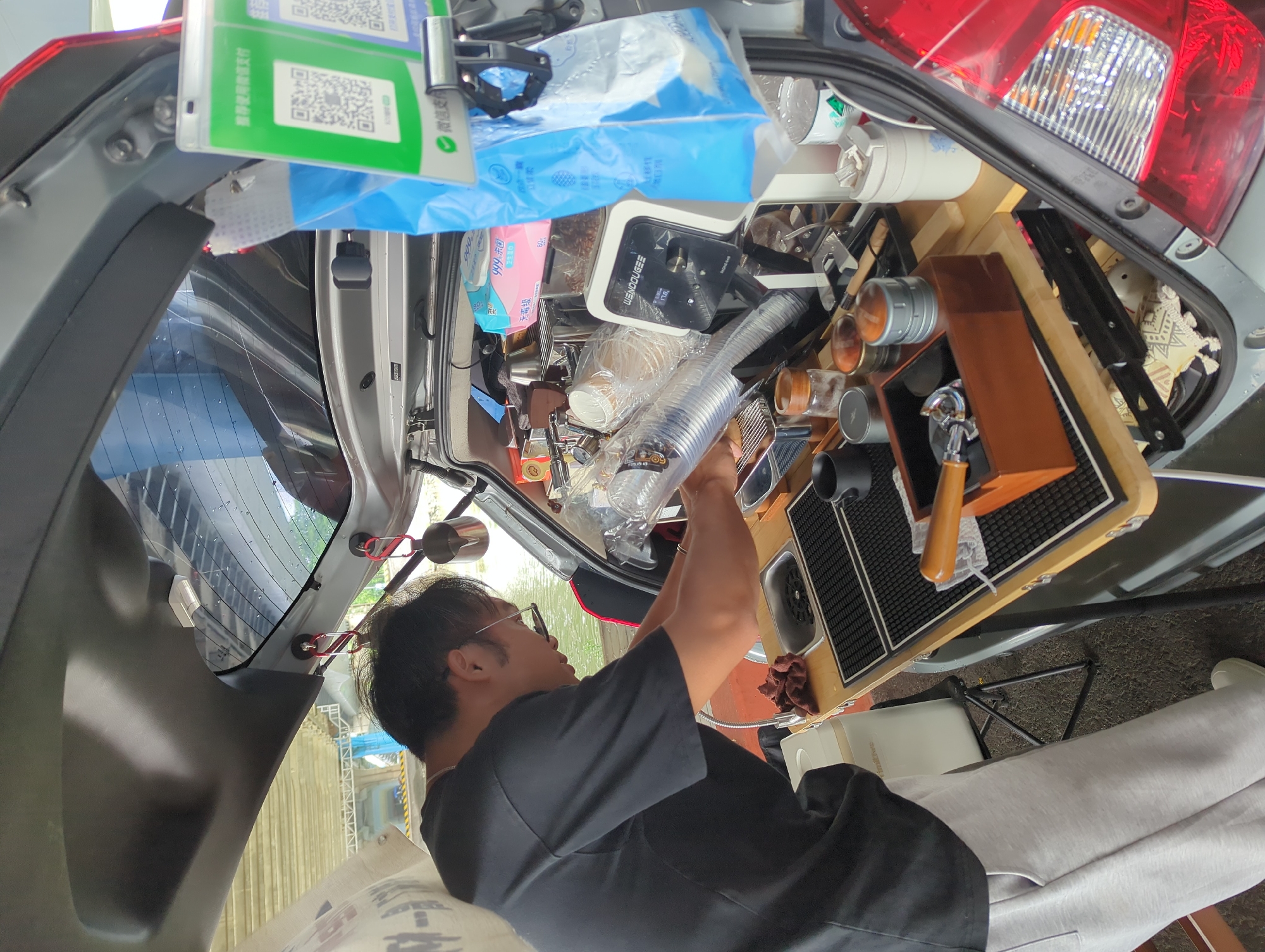 Here's a detailed description of the image:
**Scene:** The image appears to be taken from inside the open trunk of a car, converted into a mobile coffee station. The trunk is densely packed with coffee-making equipment and supplies.
**People:** A man is present, seemingly operating or organizing the coffee station setup. He is wearing glasses and a black t-shirt. He's turned towards the back of the car, working on the equipment.
**Objects:**
* Various coffee-related items: espresso machine parts (portafilters, tampers), coffee beans, grinders, cups, containers, and cleaning supplies.
* Electronic equipment: A small electronic scale (marked "WENDOUGEE" with a reading of "17.8")
* Food wrappers (with Chinese characters)
* A sign with a QR code for WeChat Pay (recommended payment method indicated)
**Environment:**
* The trunk of the car is adapted with a wooden countertop to hold the coffee equipment.
* Outside the car, there are glimpses of an outdoor setting - possibly near a body of water or a park-like area.
* The scene is well-lit, appearing to be during the daytime.
**Location:** Given the use of WeChat Pay and the presence of Chinese characters on the packaging, it's likely the photo was taken in China. Additionally, the OCR output suggests that the hygiene product shown is from "Shunde District".