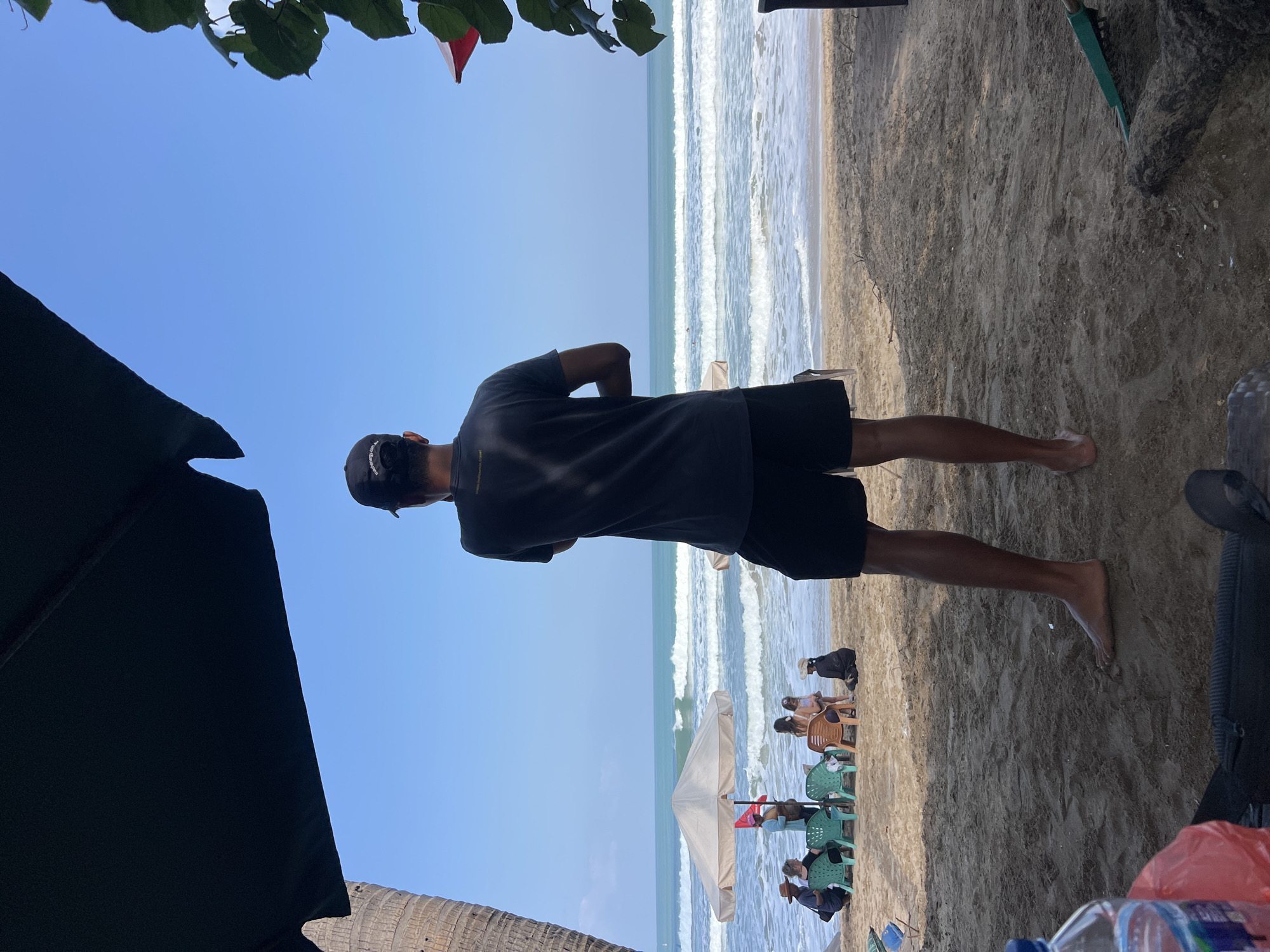 A man stands with his back to the camera on a sandy beach in Kuta, Indonesia. He is wearing a dark t-shirt, dark shorts, and a baseball cap. In front of him, several people are seated in chairs under a large umbrella. The ocean is visible in the background, with waves rolling towards the shore. The sky is clear and blue. There are palm trees and foliage framing the scene, along with some beach furniture and a plastic bottle in the foreground.