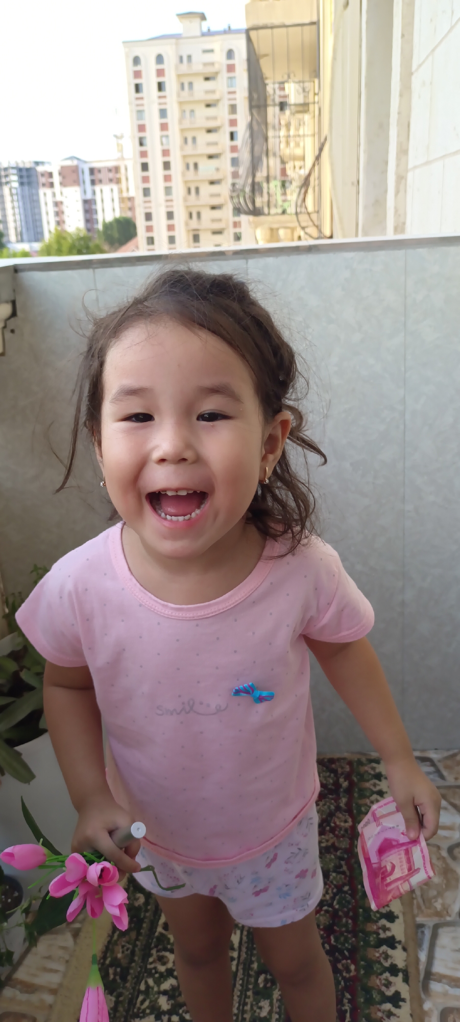 A young girl with brown hair and a pink t-shirt with a "smile" graphic and a blue butterfly clip stands on a balcony. She is holding a small bunch of pink flowers in her left hand and a pink item that resembles money in her right hand. The background shows buildings, trees, and a sky typical of Fergana City, Uzbekistan. The girl is smiling broadly, showing her teeth, and appears to be mid-laugh. The balcony railing is visible in the foreground, and a patterned rug is on the floor. The lighting suggests it is daytime.
