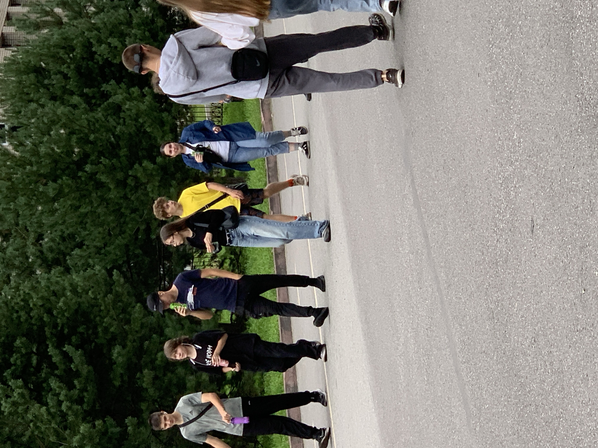 A group of seven young people, appearing to be teenagers or young adults, are walking on an asphalt road next to a line of green trees. Most of the individuals are casually dressed in t-shirts, jeans, and athletic wear. One person on the far left is wearing a grey t-shirt and black pants, holding a purple water bottle. Next to them, a person in a black t-shirt and black pants has a drink in their hand. To their right, a person in a blue t-shirt and black pants is drinking from a green can. Following them, a person in a yellow t-shirt and dark shorts is walking. Further down the line, a person in a black jacket and blue jeans is looking ahead. Another person, in a blue jacket and blue jeans, is also walking with the group. The person on the far right, wearing a grey hoodie and grey pants, has a black crossbody bag. They are all walking in the same direction, appearing to be outdoors on a cloudy day. The environment suggests a suburban or park-like setting within the city of Prokopyevsk, Russia.