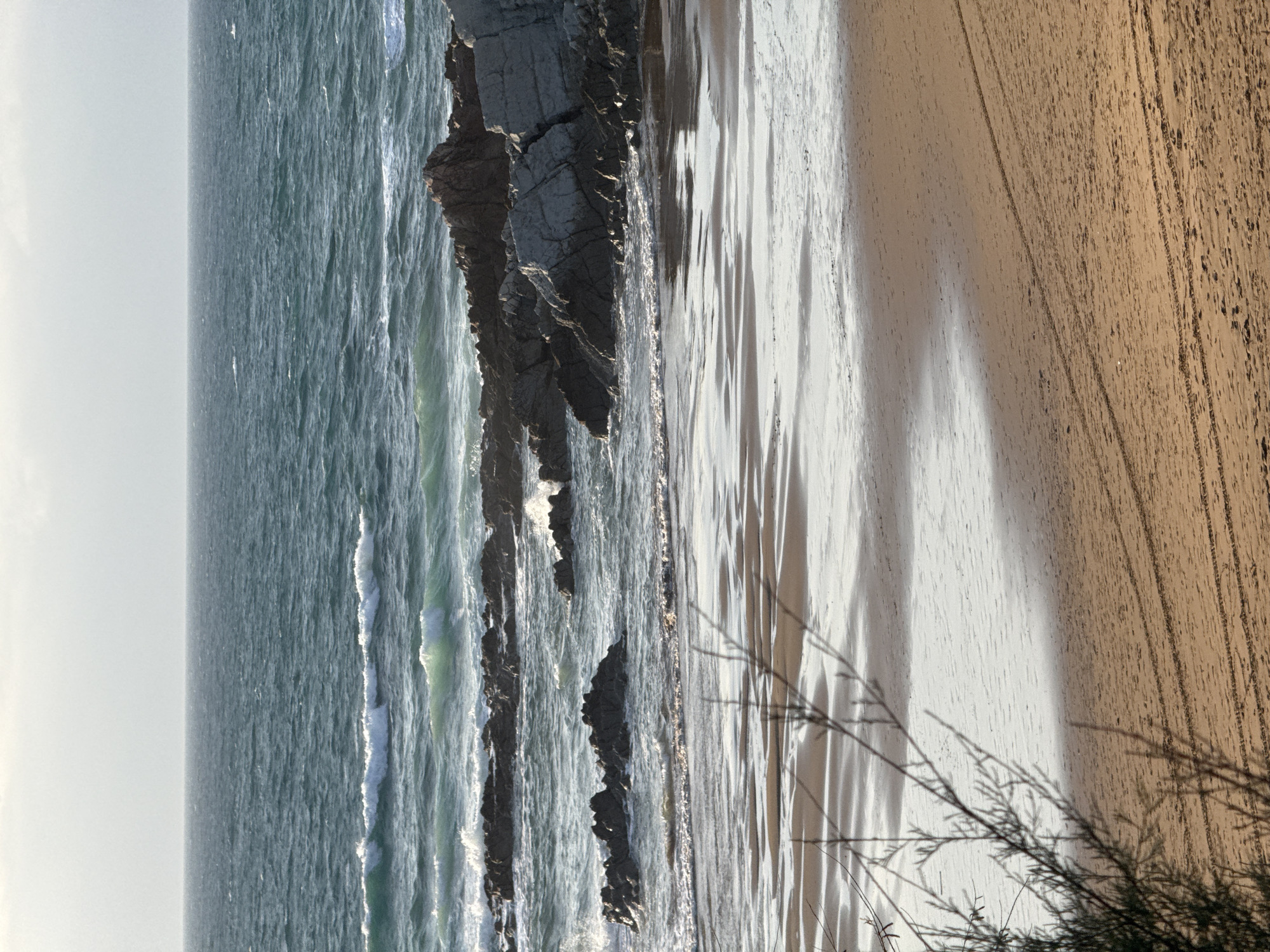The image shows a beach scene in Sopela, Spain. The foreground features wet sand with tire tracks. In the mid-ground, the shallow water reflects the sky and the darker rocks and the sea. In the background, the choppy, deep teal ocean stretches to the horizon under a pale sky. Rocky outcrops emerge from the water, with waves crashing against them. A bare branch of a bush is visible in the lower left corner.