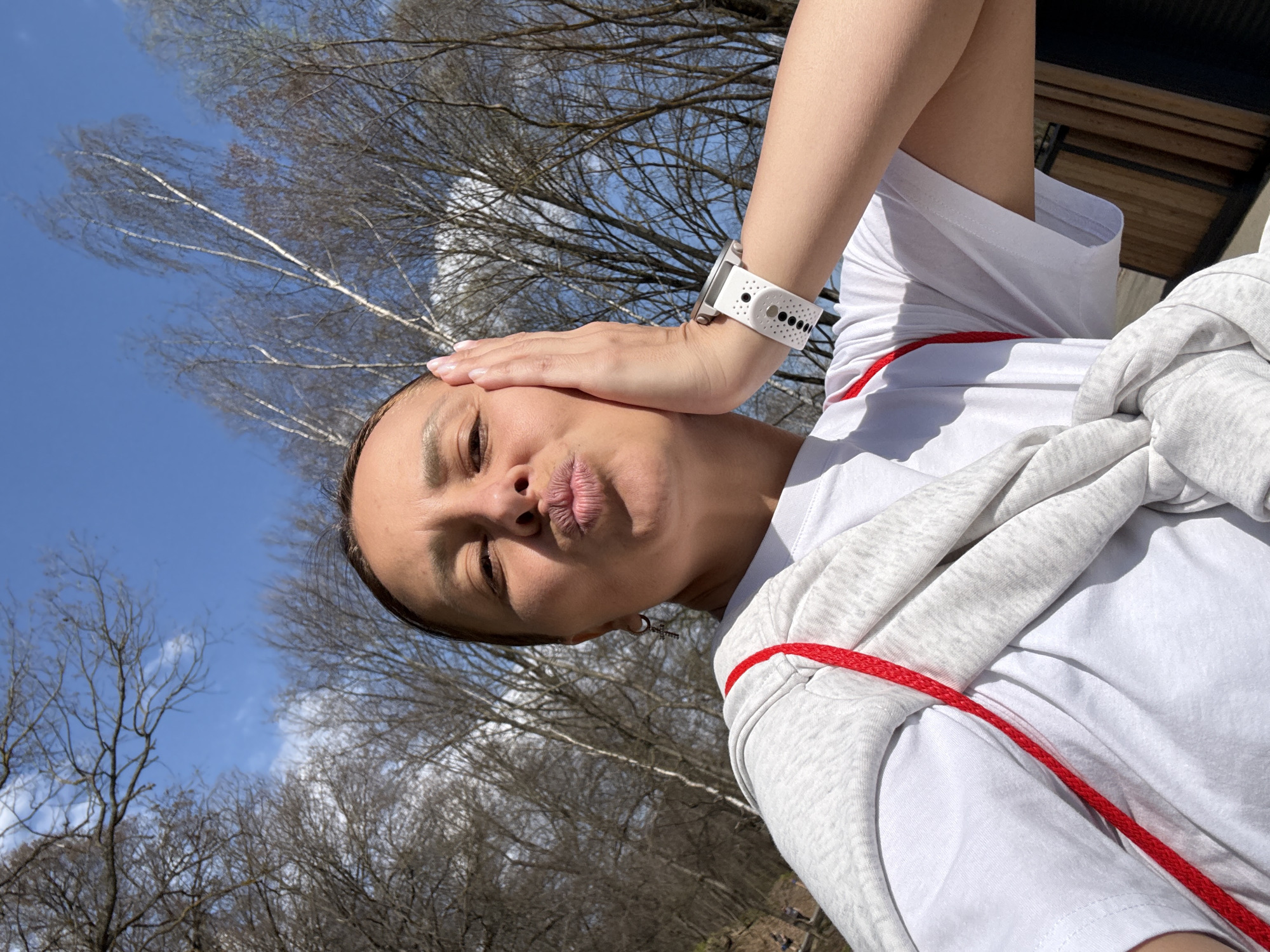 A selfie of a woman with a neutral expression, wearing a white t-shirt and a gray hoodie draped over her shoulders with a red drawstring visible. Her right hand is touching her forehead, and she's wearing a white smartwatch. The background shows bare trees against a blue sky with sparse clouds. The location is consistent with an outdoor setting in Sdt Shcherbakovka, Russia, given the trees and sky, but there is insufficient detail to be certain.