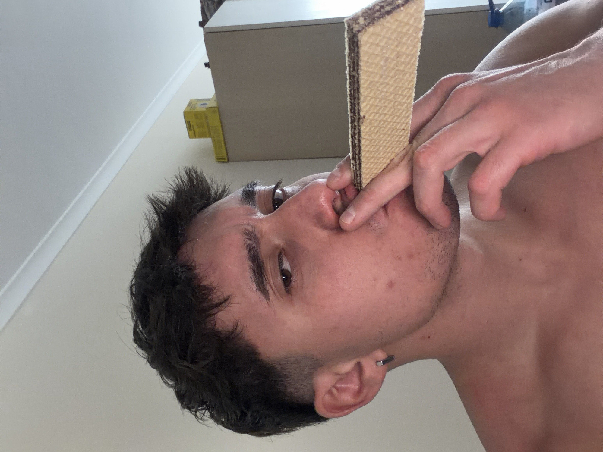 A shirtless man with dark hair is positioned in the foreground with a wafer biscuit held against his nose. His left ear is visible, adorned with a silver hoop earring. His left hand is holding the biscuit near his nose. The background contains a beige cabinet with an open container on top. A yellow box is visible behind the cabinet. The environment suggests an indoor setting, likely a residence. There is no evidence of the city of Sofia, Bulgaria.
