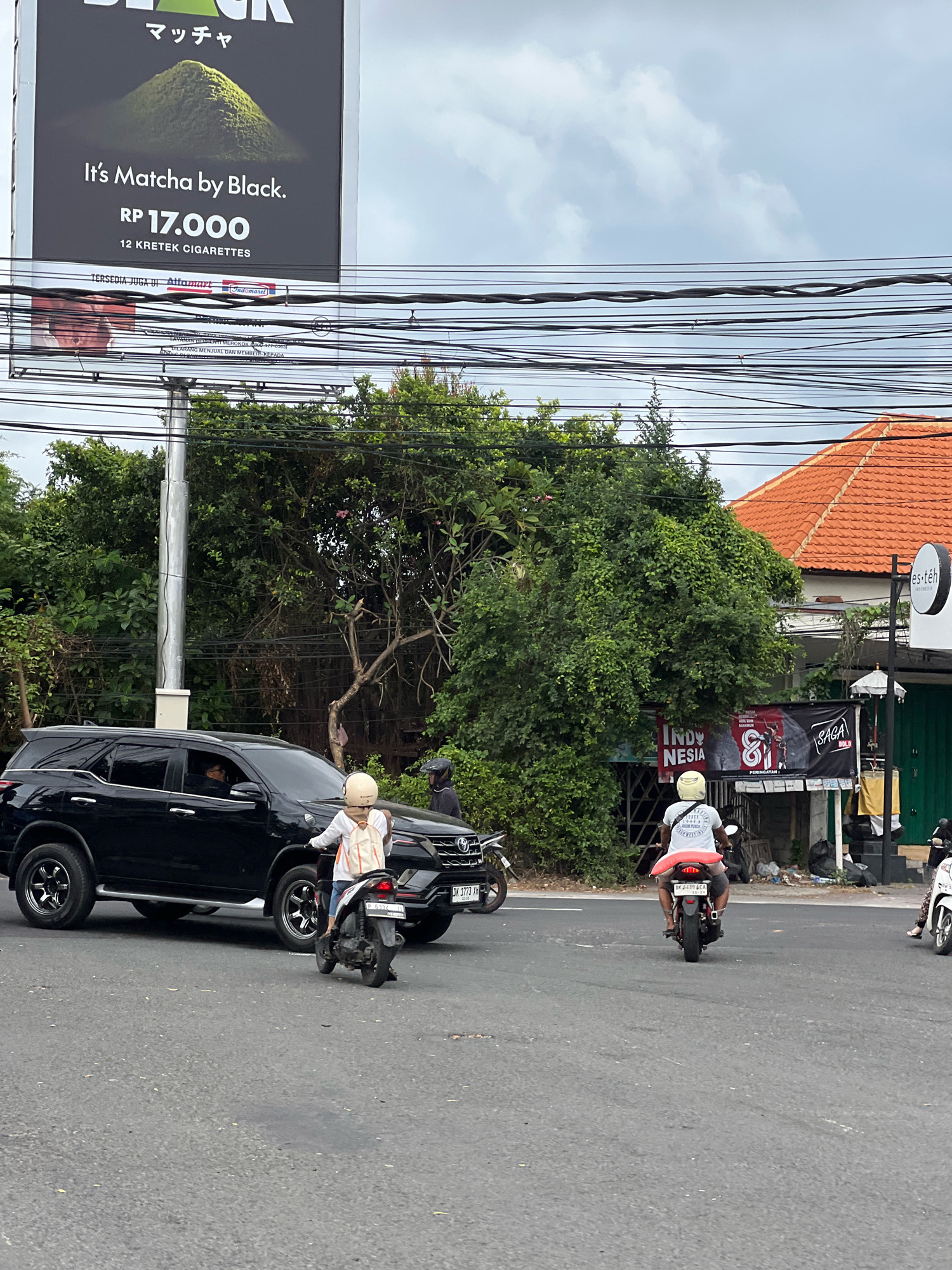In Denpasar, Indonesia, on a cloudy day, a black SUV is stopped at an intersection with its driver visible behind the wheel. A person wearing a white shirt, light-colored helmet, and a backpack is riding a scooter in front of the SUV. To the right, another person on a scooter with a white helmet is also at the intersection. In the background, a building with an orange tiled roof and a sign that reads "Es Teh" is visible. A large billboard advertising "It's Matcha by Black" cigarettes, priced at Rp 17,000 for 12 kretek cigarettes, is prominently displayed. Various other banners and signs are also visible in the background, along with dense green foliage and utility wires overhead.