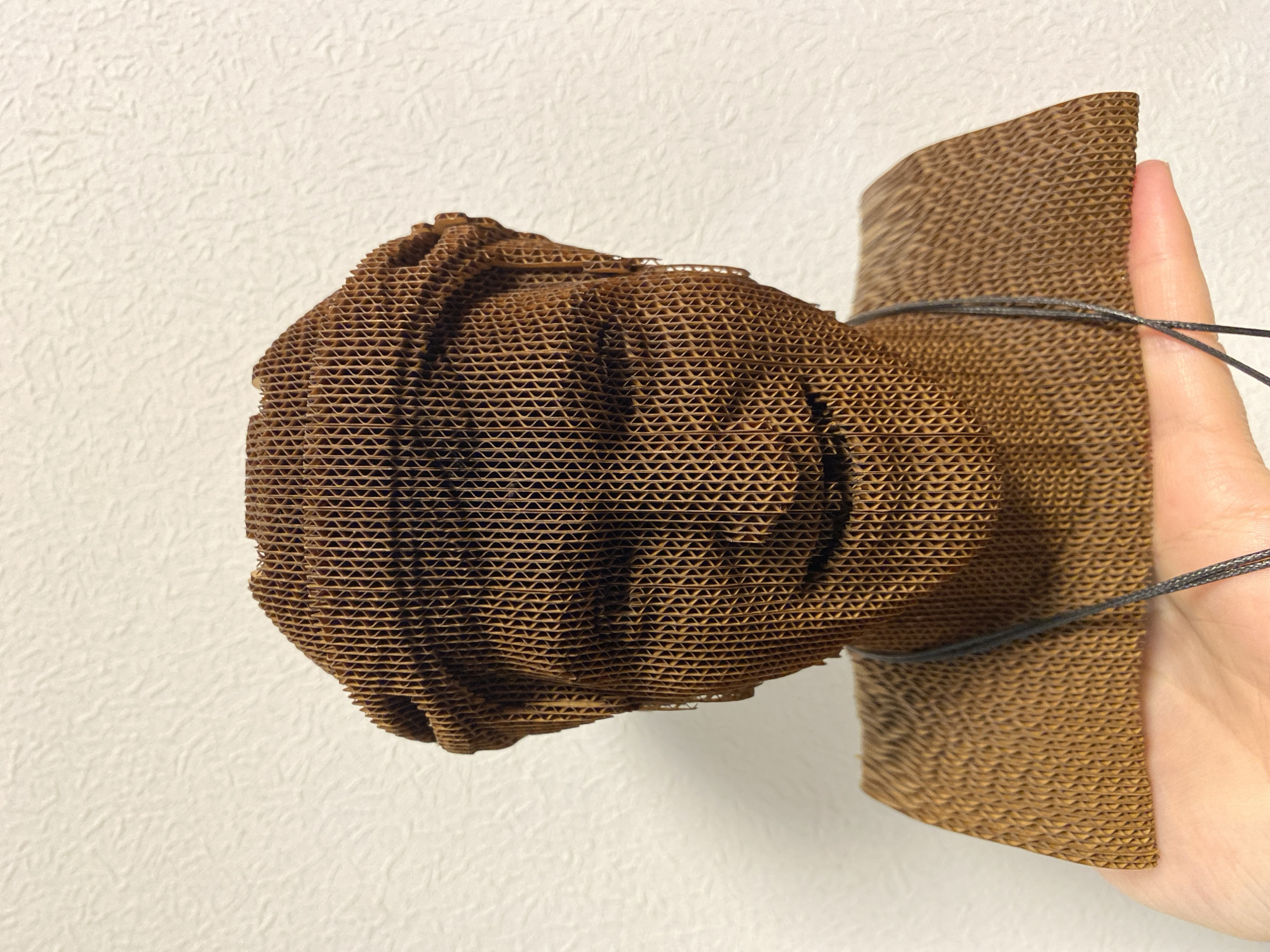 A hand holds a brown, corrugated cardboard art piece against a white wall. The art piece consists of a crumpled, rounded shape attached to a more cylindrical, upright piece. A thin, dark string is wrapped around the upright section and a finger. The wall is textured. The location in Chaikovsky, Russia is not known from this image.