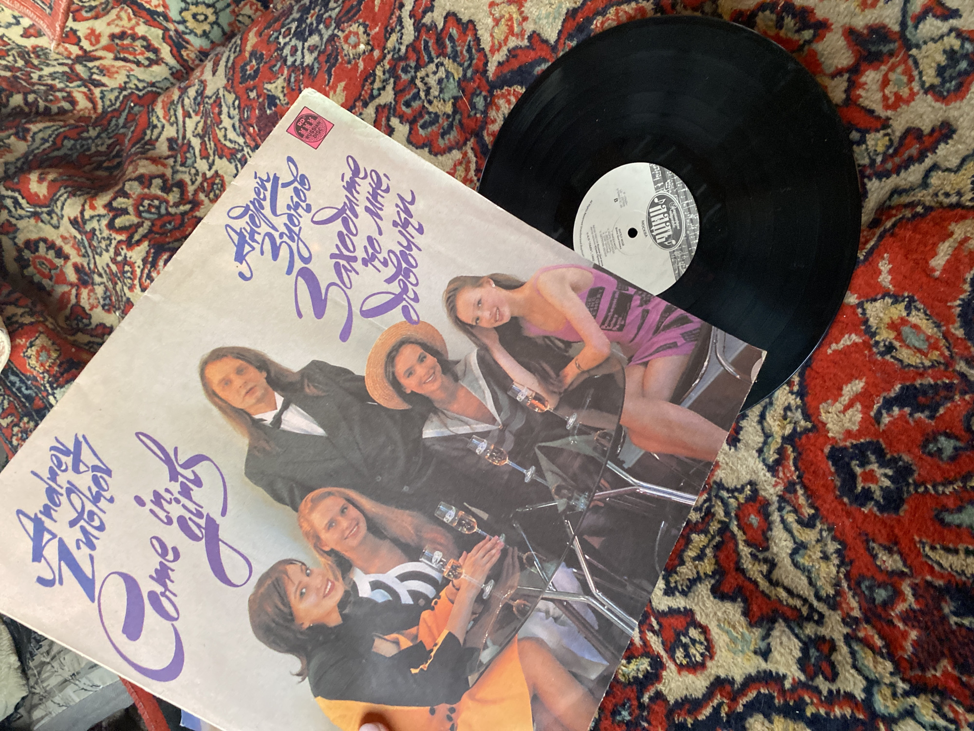 The image shows a vinyl record and its album cover resting on a patterned rug. The album cover features a group of people, including a man in a tuxedo and several women, sitting around a glass table with champagne glasses. The album title is "Andrey Zlotkov Come in girls" (written as "Авдрей Злотков Заходите ко мне, девушки" in Cyrillic). The record is black with a white label. There is no indication of the image being in the city of Ольховка, Russia.