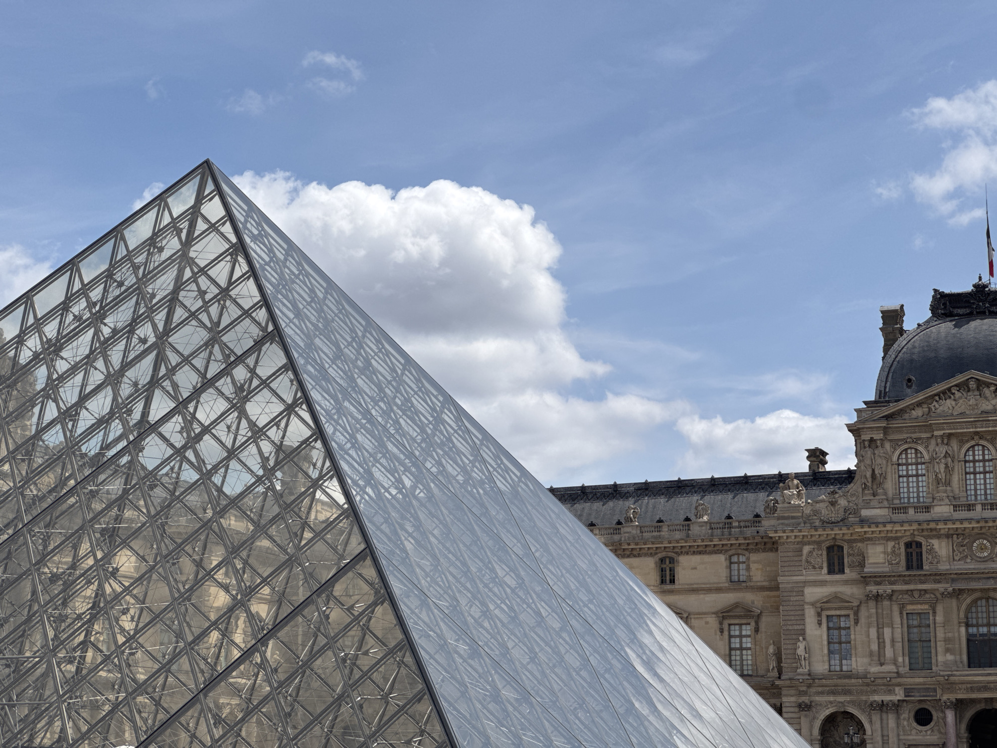 The image shows a close-up view of the Louvre Pyramid in Paris, France. The modern glass pyramid stands in stark contrast to the historic architecture of the Louvre Museum in the background. The pyramid's intricate geometric structure is visible, reflecting the sky and the surrounding buildings. The sky is blue with scattered white clouds. The Louvre Museum's facade features classical architectural elements, including ornate carvings, arched windows, and a domed roof. A French flag is visible atop one of the museum's structures. There are no people visible in the image. The scene is a depiction of the architectural juxtaposition of old and new in a prominent Parisian landmark.