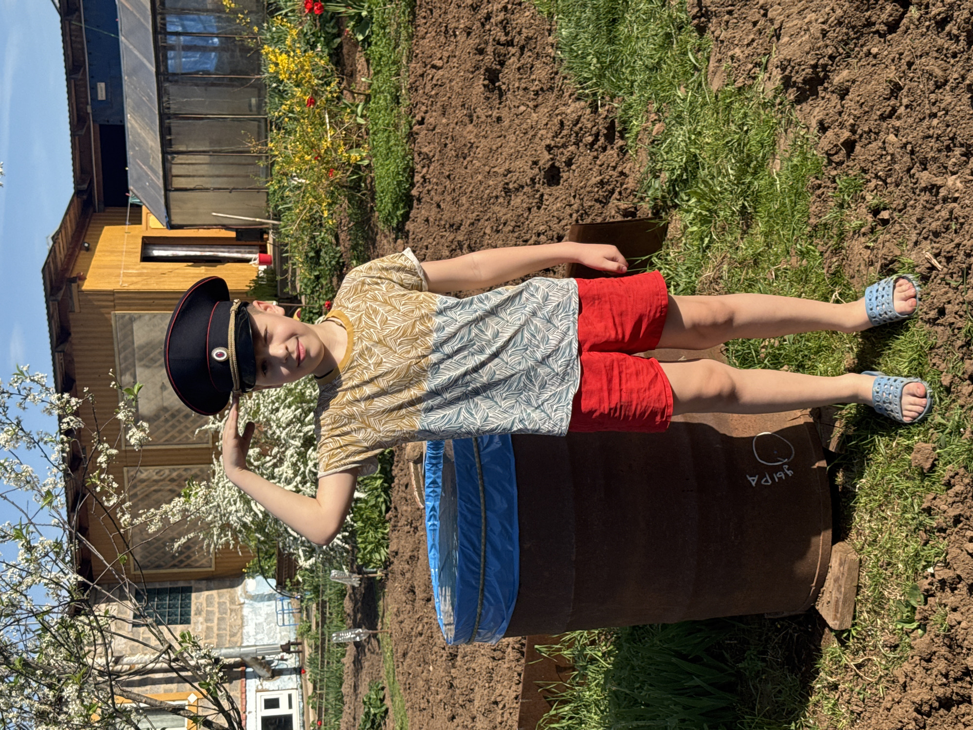 A young male child is standing in a garden in what appears to be the city of Dubovaya, Russia, based on the Cyrillic script visible on a rusty barrel next to him. He is wearing a military-style peaked cap, a patterned short-sleeved t-shirt, red shorts, and blue sandals. He is saluting with his right hand. The garden is a mix of cultivated soil and grass. In the background, there is a wooden structure resembling a greenhouse and what appears to be a house, as well as blossoming trees and other plants. The weather appears sunny, based on the shadows and clear sky.