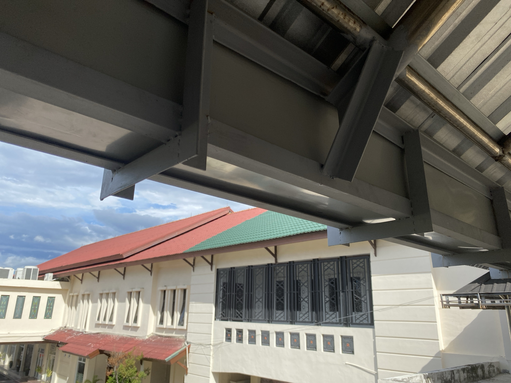 An exterior shot captures a building in Banda Aceh, Indonesia, with a focus on its roofline and architectural details. The building features a red-tiled roof, with a section of green tiles visible as well. The walls are painted white and have a series of windows, some with decorative dark-colored grilles. Above the building, a metallic overhang with structural beams is prominent, extending into the foreground of the image. The sky above is partly cloudy, with a mix of blue sky and white clouds. The scene appears to be taken from an elevated vantage point, looking downwards towards the building and its surroundings.