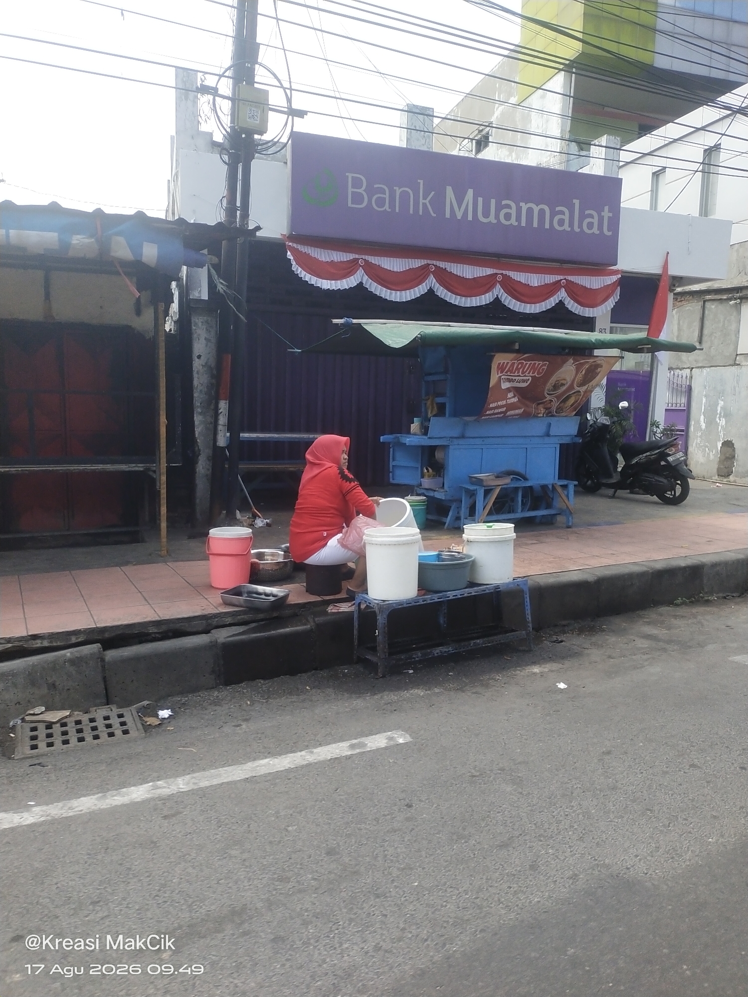 The image depicts an outdoor scene in the city of Kalijaten, Indonesia, likely on a sidewalk next to a street. In the foreground, a person wearing a red hijab and white pants is sitting on a dark stool, engaged in an activity that appears to involve washing dishes or food preparation. Several white and pink buckets, a metal basin, and a rectangular metal tray are arranged around the person on the tiled sidewalk. To the right of the person is a blue food cart or stall with a green awning, displaying a banner that reads "WARUNG" with images of food. A black motorcycle is parked near the food stall. Behind the stall, a building with a purple facade bears the sign "Bank Muamalat". Overhead, numerous electrical wires crisscross the sky. The street in the lower portion of the image shows a gray asphalt surface with a white line marking. The overall atmosphere suggests a commercial or street-side food service operation in a moderately urban environment. The image is watermarked with "@Kreasi MakCik 17 Agu 2026 09.49".