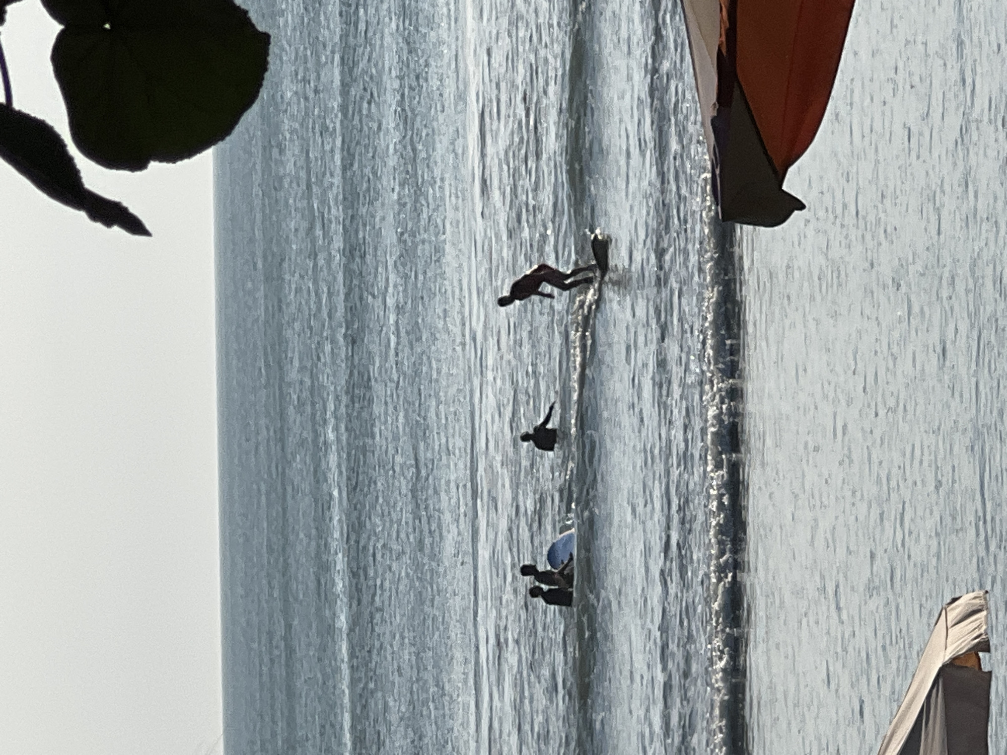 In Kuta, Indonesia, at least four individuals are visible in the ocean. One person is actively surfing on a surfboard, appearing to be mid-ride. Another group of three individuals are in the water, some appearing to be sitting or kneeling on surfboards. The environment is a beach scene with the ocean taking up the majority of the frame. The sky is overcast, and there is a hint of foliage from a tree in the upper right corner. The bottom of the image shows a portion of what appears to be an umbrella or beach canopy. The water is calm with small waves breaking in the distance.