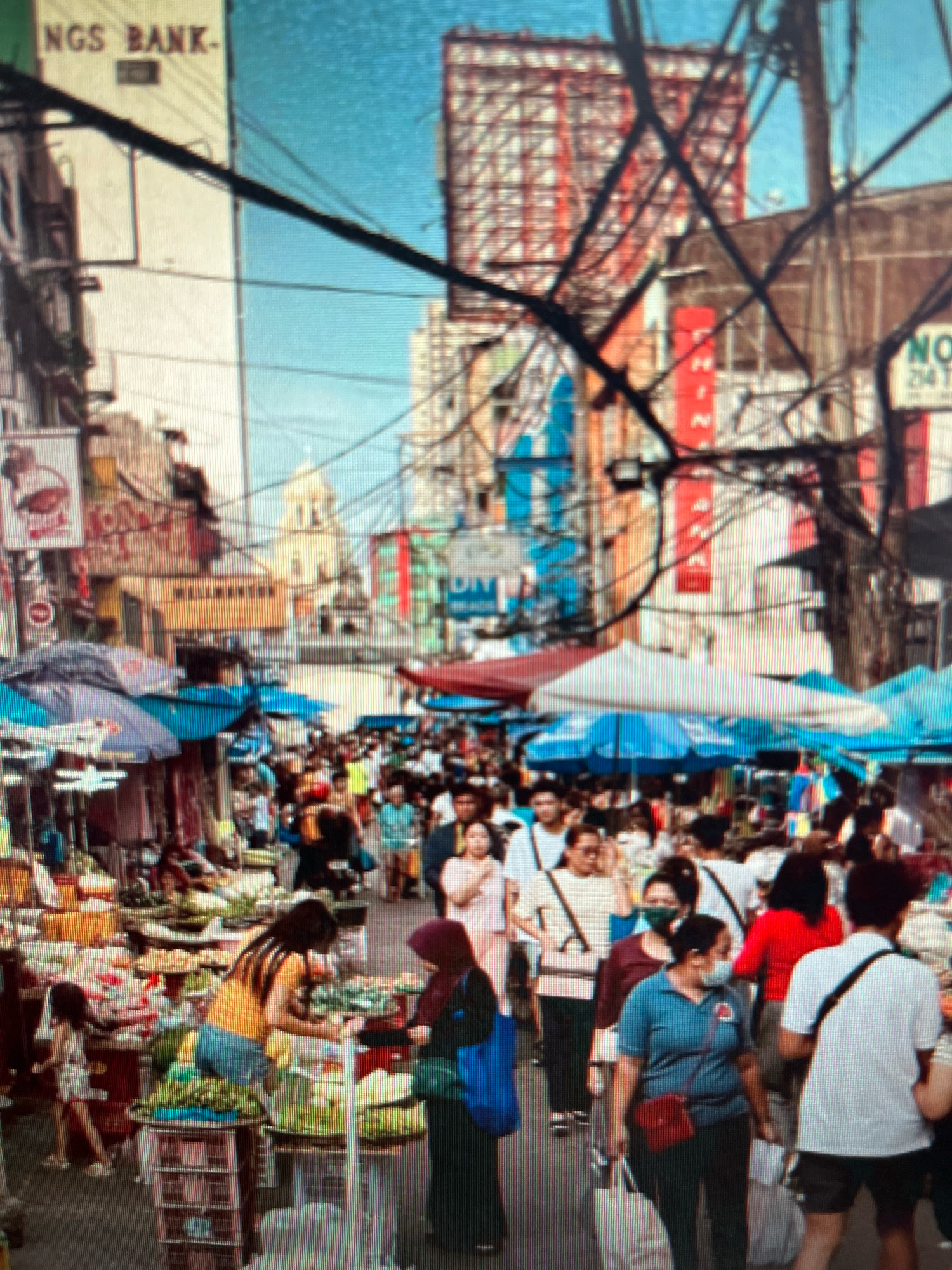 This image depicts a crowded street market in Sejahtera, Indonesia. The scene is bustling with activity, featuring a dense congregation of people navigating narrow pathways lined with market stalls. Several individuals are seen interacting, including vendors attending to their goods and shoppers browsing.
The market stalls display a variety of merchandise, with an emphasis on fresh produce like vegetables and fruits. Colorful umbrellas and canopies are set up to provide shade for the vendors and their products. In the background, buildings with signage and architectural details indicate an urban setting. The overall atmosphere is lively and dynamic, characteristic of a busy marketplace in a Southeast Asian city.