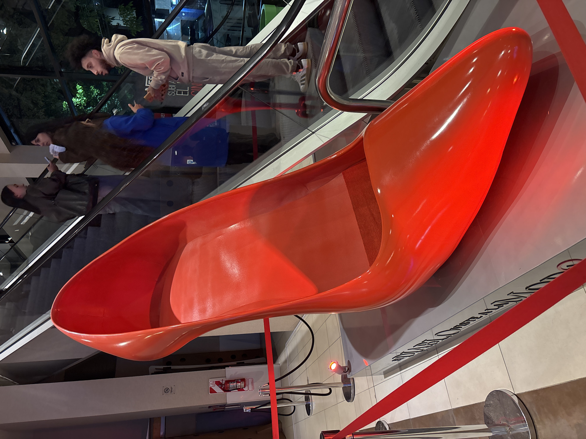 This image, taken in Buenos Aires, Argentina, captures an indoor scene dominated by a large, vibrant orange high-heeled shoe sculpture or chair. The shoe, a glossy, exaggerated pump design, is positioned on a raised platform, illuminated by a small red spotlight, and cordoned off by red velvet ropes and chrome stanchions, suggesting it is an exhibit or art installation.
In the background and reflected in the glass barriers, several people are visible riding an escalator. A young man with dark, curly hair and a beard, wearing a light-colored tracksuit, is looking downwards, possibly at his phone. Behind him, another person with long, dark hair, dressed in a brown jacket over a blue garment, also appears to be using a phone. Further down the escalator, a third individual in a dark jacket is also present.
The setting appears to be a modern commercial building or mall, characterized by the escalators with glass side panels and artificial lighting. The overall ambiance suggests an evening or nighttime interior. Notable details include a fire extinguisher mounted on a pillar behind the roped-off area.
Visible text includes partial words on a sign behind the escalators, such as "SUBI" and "EL." On the floor/wall behind the shoe, obscured by the rope, faint black cursive text is visible, which is difficult to decipher fully.