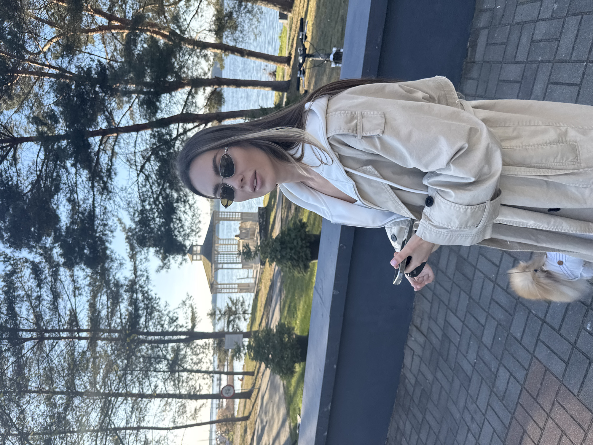 A woman with long blonde hair wearing sunglasses and a beige trench coat stands on a brick-paved surface. She is holding a phone in her right hand, and a dog, primarily the tail of which is visible, is present at her side. Behind her is a low concrete wall, and beyond that, a park with trees and a body of water are visible. In the background, there is a structure resembling a gazebo or small pavilion.
The location appears to be a park near water in Białobrzegi, Poland, as indicated by the presence of typical Polish city features. The photo captures an everyday scene of a woman with her dog in a public park.