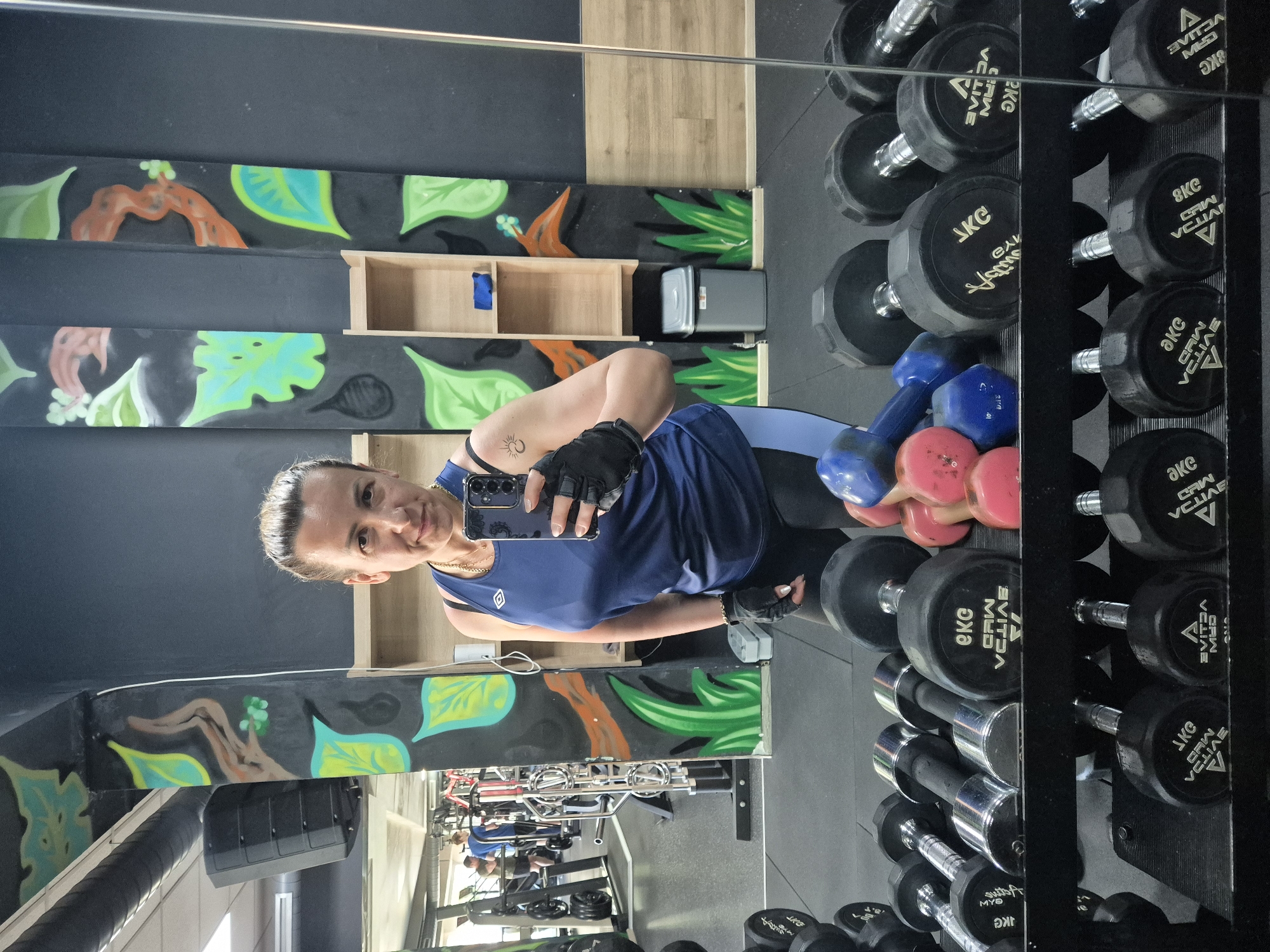 A woman with brown hair pulled back, wearing a blue tank top and black leggings, takes a selfie in a gym. She is holding a black smartphone in her right hand and wearing black gloves. In the background, a wall with colorful jungle-themed murals is visible. Shelves with small items and a trash can are also present. In the foreground, rows of dumbbells are neatly arranged on racks. The woman has a sun tattoo on her left arm. The gym appears to be well-equipped with various exercise machines visible in the reflection of a mirror.