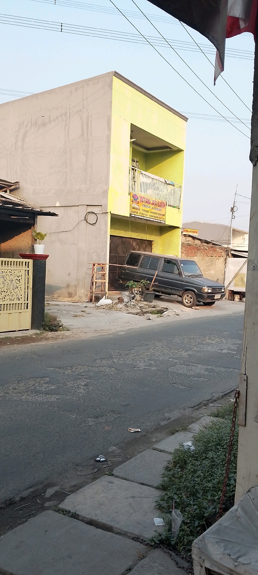 The image depicts a street scene in Satriajaya, Indonesia. In the foreground, a gray asphalt road with visible damage and scattered debris leads towards a sidewalk made of concrete slabs. Adjacent to the sidewalk is a patch of dense green vegetation, partially obscured by a rusty chain.
Dominating the midground is a two-story building with a pale gray first floor and a bright lime green second floor. The second floor features a balcony with a railing. A black van is parked in front of the building's dark brown garage doors. To the left of the building, a yellow wrought-iron gate stands beside a dark gray pillar with a potted plant. A wooden ladder leans against the side of the building, near a pile of white material and other assorted debris. A banner with red, white, and blue accents hangs on the lime green facade, displaying text in Indonesian, including the name "MUTIARA JAYA PLASTIK."
In the background, other buildings and structures are visible, including a brick wall and a white building. Power lines crisscross the pale blue sky. The overall scene suggests a commercial or residential area in an Indonesian town, characterized by a mix of new and aging infrastructure, and evidence of everyday street life with scattered refuse.