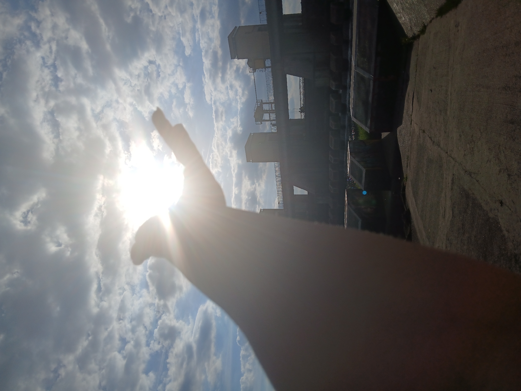 The photograph depicts a sky with clouds and the sun, partially obscured by a human hand reaching towards it. In the background, there is a structure resembling a dam or industrial building. The hand is blurred, likely due to being close to the camera lens. The image is taken in the city of Unknown, Unknown. The picture looks up at the sky with clouds and sun.