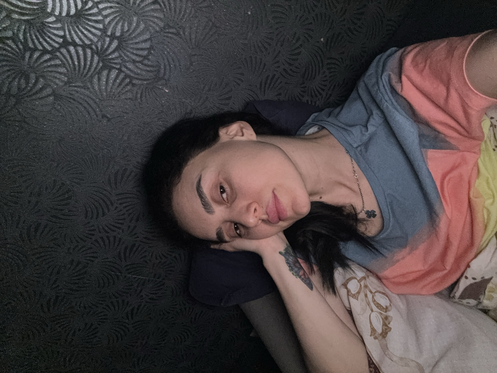A person is reclining on a patterned surface with a pillow or cushion, resting their head on their hand. The individual has dark hair, prominent eyebrows, and a visible tattoo on their forearm. They are wearing a multicolored t-shirt and a necklace. The background consists of a textured wall with a repeating, stylized leaf or petal pattern. The location cannot be ascertained from the image.