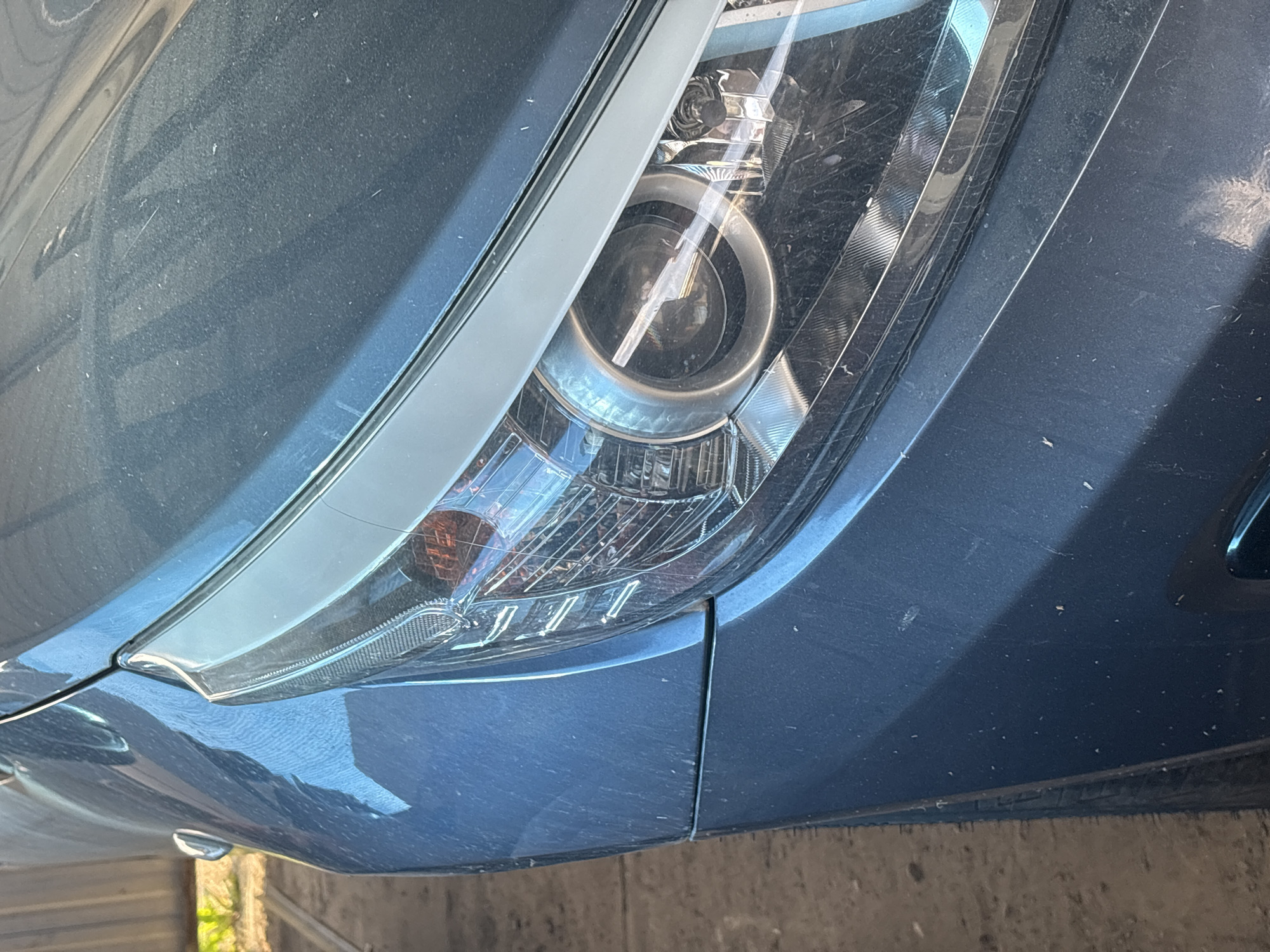 This media file is a close-up, low-angle shot of the front right headlight and fender of a dark blue car. The car appears to be dusty, with visible specks of dirt and some minor scratches on the surface, particularly on the fender. The headlight itself is complex, featuring a projector lens with metallic accents and a clear, ribbed lens cover. An amber turn signal is visible within the headlight assembly. The car is positioned outdoors, as suggested by the dappled sunlight reflecting off the metallic paint and the presence of what appears to be vegetation and a weathered wooden fence in the background, indicating a rural or semi-rural setting. The lighting suggests it is daytime, possibly a sunny day due to the bright reflections. No people are visible in the frame. The overall impression is of a vehicle that has been used and exposed to the elements. The metadata indicates the location is Sovkhoza "Razdol'e", Russia. There is no visible text in the image.