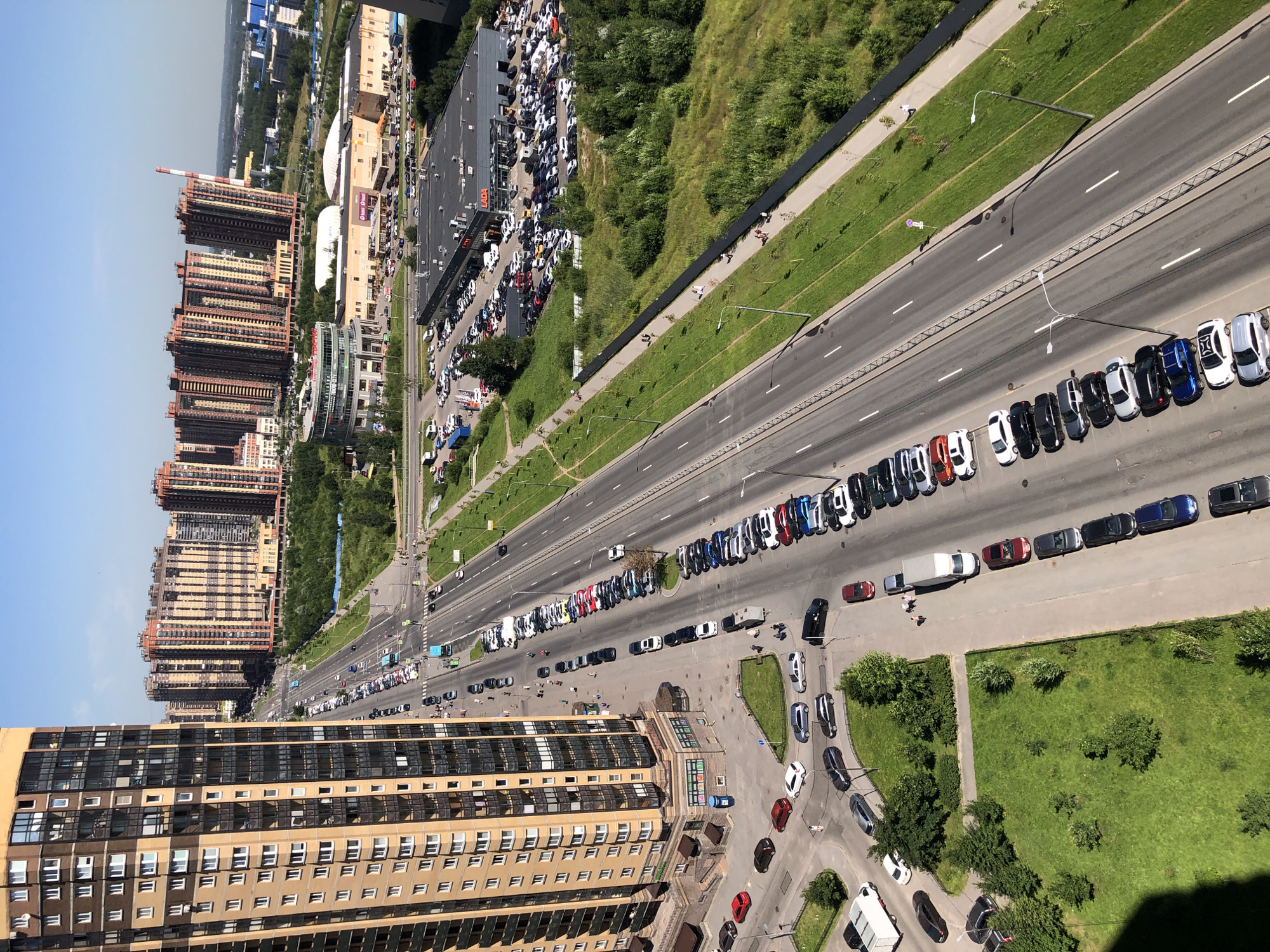 The image is an aerial view of a city street with buildings and parked cars. On the left, a tall, modern apartment building with a tan and brown facade dominates the foreground. Across the street, a multi-lane road is visible with cars parked on the right side. In the background, several high-rise residential buildings with red and beige exteriors are clustered together. Further in the distance, commercial buildings and a power plant smokestack are visible. The scene is set on a sunny day with a clear blue sky.
