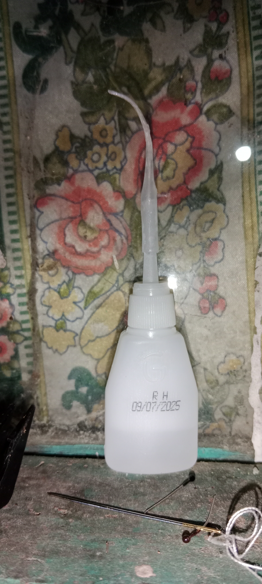 In Subang, Indonesia, a white bottle of adhesive is positioned on a surface with a floral pattern. The bottle has a slender applicator tip and a white cap. Embossed on the bottle is a logo, possibly a stylized "G," and text indicating "RH" followed by the date "09/07/2025." In the foreground, a thin metal rod, likely a needle or probe, lies horizontally. To the right of the bottle, a few small metallic and dark-colored objects are visible, along with some white thread or string. The background features a faded floral textile. The overall scene suggests a workspace or hobby area.