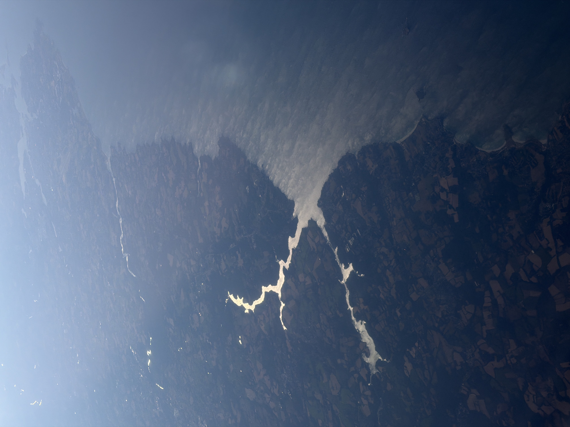 This is an aerial photograph of the coastline near Fouesnant, France, with a body of water on the right side of the image and land on the left. The land appears to be a mix of agricultural fields and forested areas, with some settlements visible. The coastline is irregular, with bays and inlets. The water is a deep blue, and there appears to be some haze or mist over the water near the coastline. The sun is shining, causing reflections on the water and highlighting certain areas of the land. The overall impression is one of a rural, coastal landscape.