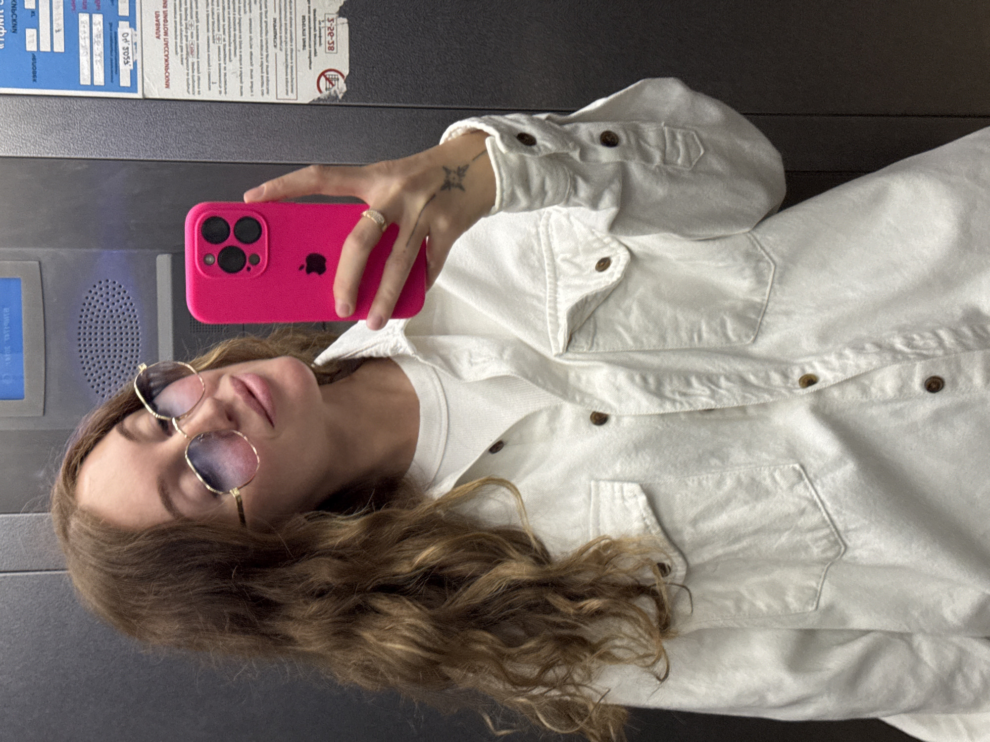 A woman is taking a selfie in what appears to be an elevator or similar enclosed space. She is holding a bright pink smartphone with three camera lenses. The woman is wearing a white, button-down jacket over a white t-shirt and gold-rimmed glasses. Her hair is long and wavy. A small tattoo is visible on the back of her hand. The background reveals the elevator's interior, including what appears to be a control panel and informational signage in Russian. The signage, along with context clues such as the environment and language on the panel, suggest the photo was taken in Chaikovsky, Russia.