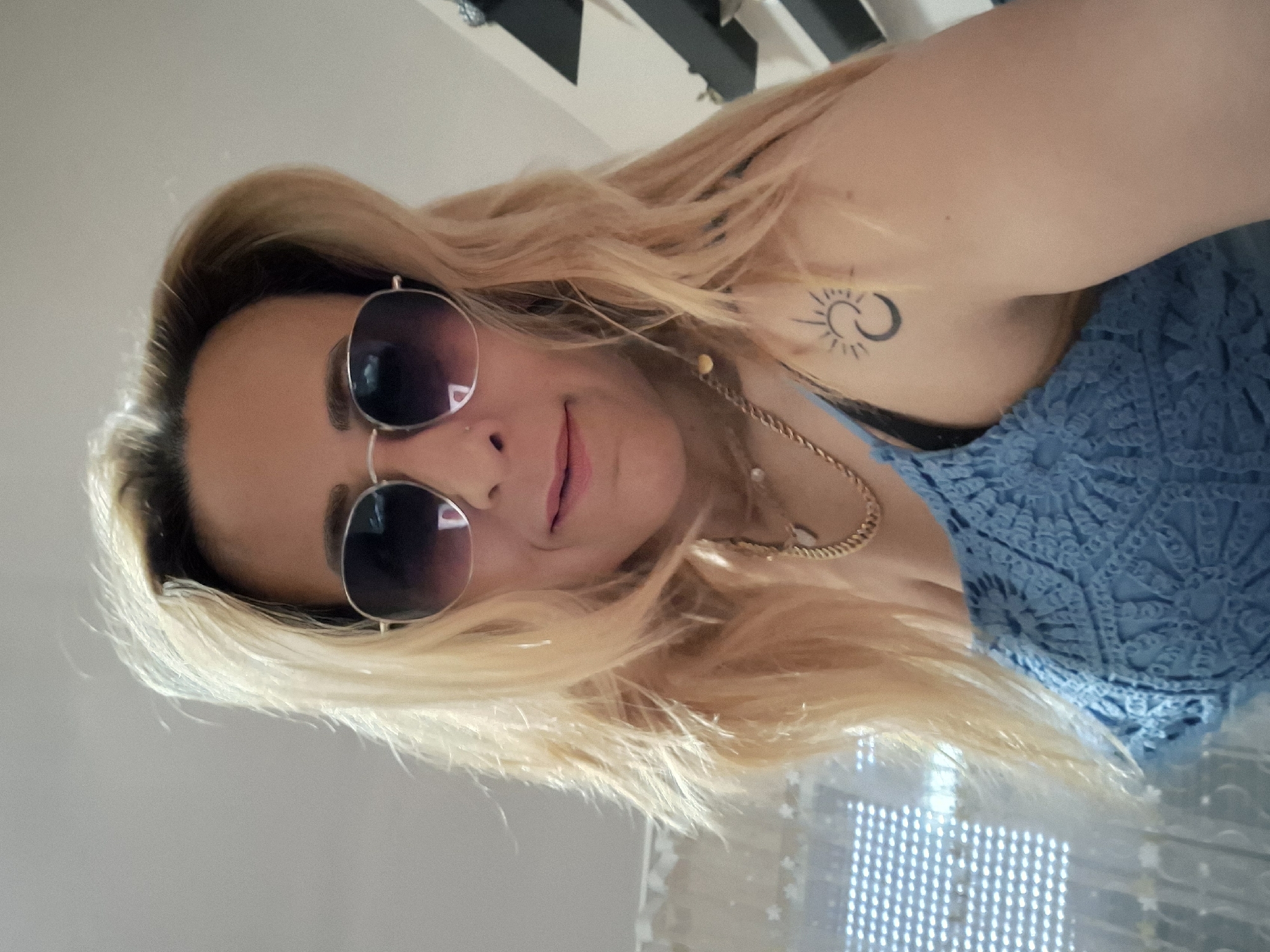 An extreme close-up, waist-up, selfie-style shot shows a blonde woman wearing sunglasses and a blue crocheted top. The woman's face is angled towards the camera, and she is smiling. She has blonde hair that falls past her shoulders. The woman is wearing a gold chain necklace and a tattoo of a sun and moon on her right shoulder. The background is blurred, but it appears to be an indoor setting with a window on the left. The lighting is bright, suggesting it is daytime.