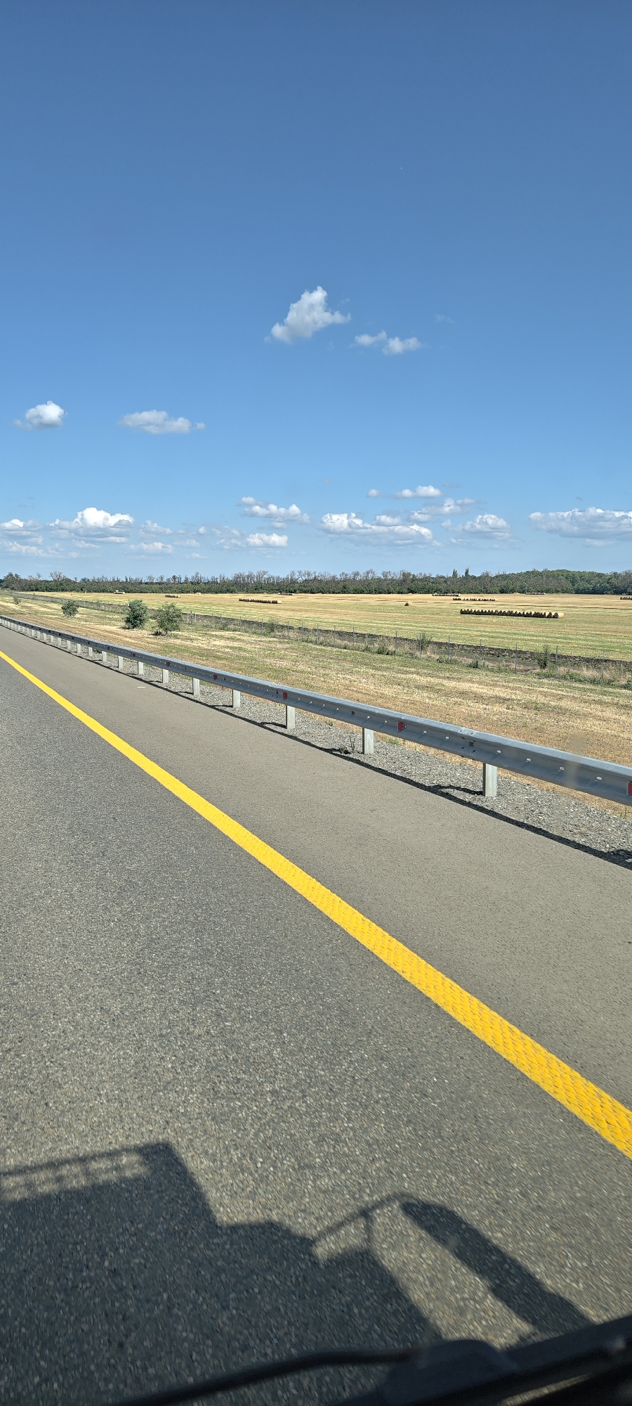 The image captures a view from a vehicle traveling on a highway, likely in the vicinity of Stavropol, Russia. The foreground is dominated by the asphalt road, marked with a prominent yellow line that guides traffic. A metallic guardrail runs along the side of the road, separating it from the adjacent landscape. Beyond the guardrail, a dry, agricultural field stretches into the distance, dotted with numerous round hay bales arranged in rows. A line of trees forms a natural border at the edge of the field, and further in the background, a hazy, treeline suggests more distant vegetation. The sky above is a clear, bright blue, with a few scattered white clouds adding depth to the scene. The overall impression is one of a rural highway on a sunny day, with agricultural activity evident in the harvested fields. The shadow of the vehicle from which the photo is taken is visible in the lower part of the image.
