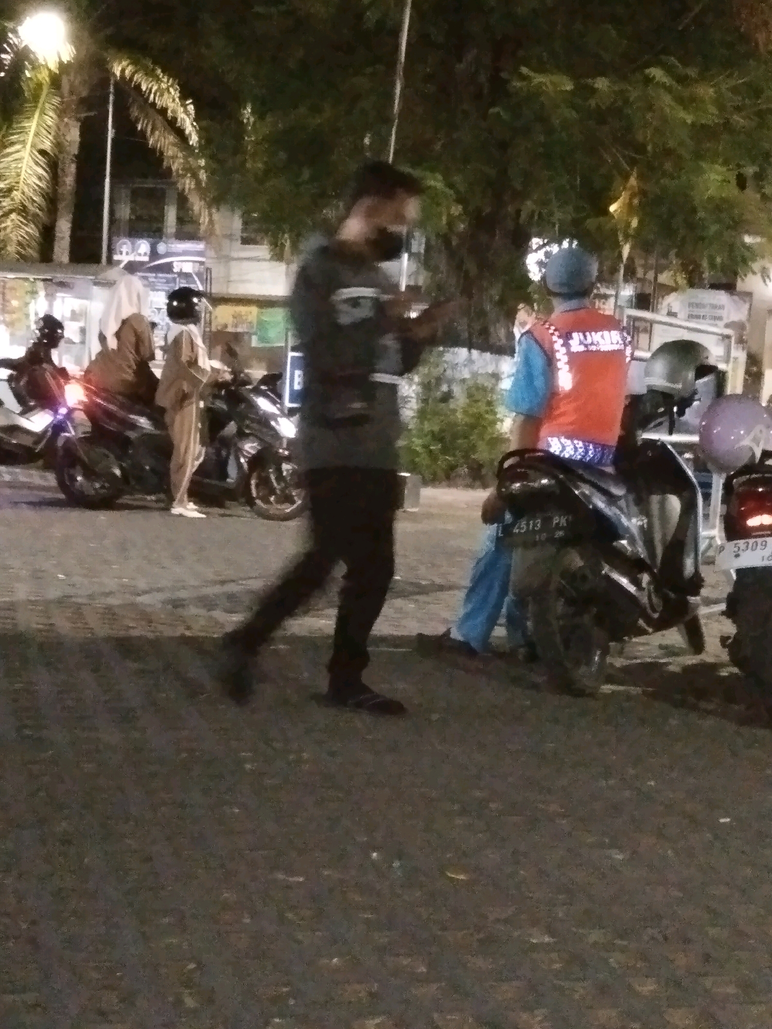 The image captures a nighttime scene in the city of Bondowoso, Indonesia. In the foreground, a person walks away from the camera, their lower body obscured by motion blur. Behind them, several motorcycles are parked. One motorcycle, in the mid-ground, displays a license plate reading "L 4513 PK." To the right of this motorcycle, another motorcycle is partially visible with a license plate that appears to read "P 5309." A person wearing an orange vest with white lettering, possibly "JUKIR," indicating a parking attendant or similar role, stands near the motorcycle with the "L 4513 PK" plate. Further in the background, two individuals on a motorcycle are visible, one wearing a hijab. Palm trees and other foliage line the background, illuminated by artificial lighting, suggesting an outdoor urban environment.