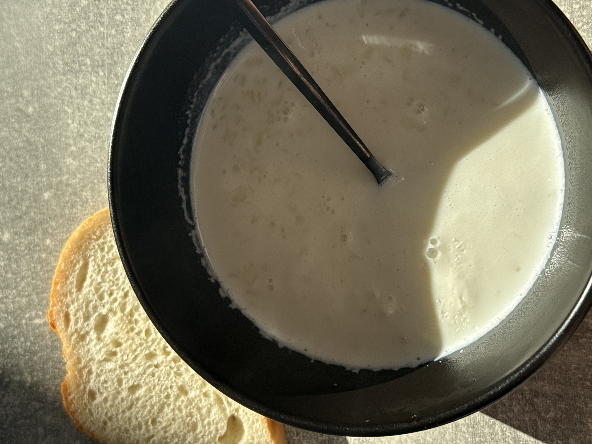 A slice of white bread is placed on a textured grey surface next to a black bowl filled with a light-colored liquid, possibly milk or a light soup. A dark spoon is partially submerged in the liquid. The scene is lit by direct sunlight, casting a shadow on the surface and the bowl. The background is out of focus. The image is taken from a top-down perspective. No information about the location in Lyubertsy, Russia, is present in the image.