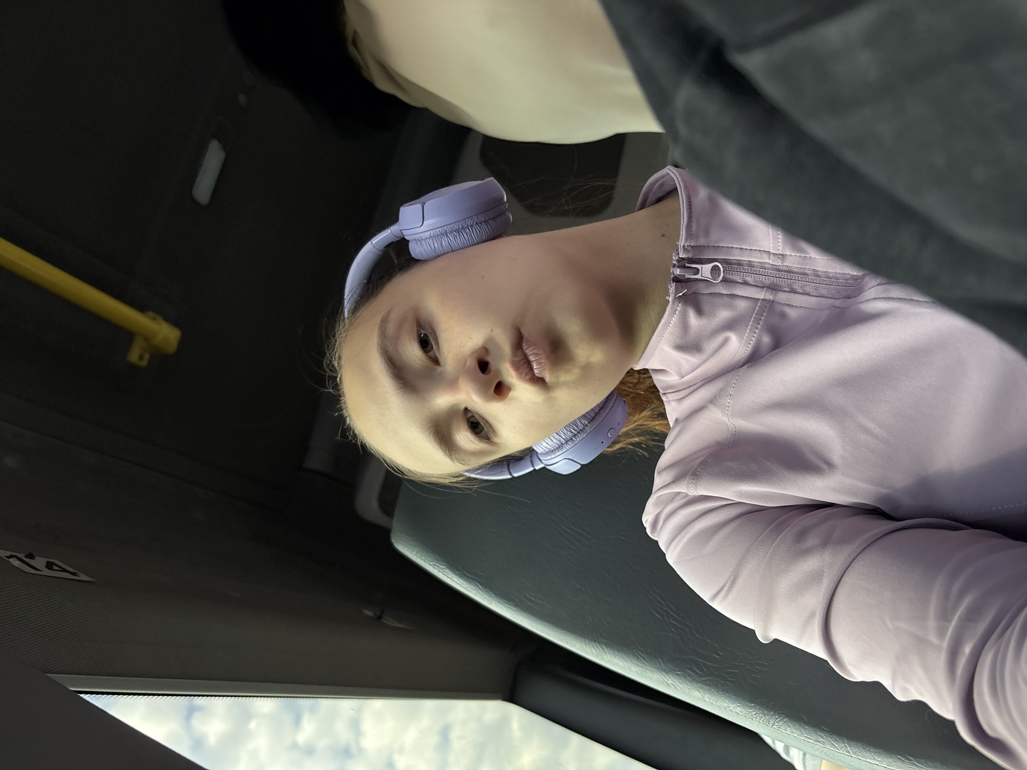 A person is seated on a bus, potentially in Черновское, Russia, evidenced by the presence of Russian text or Cyrillic characters (although not directly visible in these image crops, Черновское is assumed as context). The individual wears light purple headphones and a matching zip-up jacket. The bus interior features a gray bench-style seat and a yellow handrail. Through the window, a cloudy sky is visible. A number "14" can be seen on the bus interior.