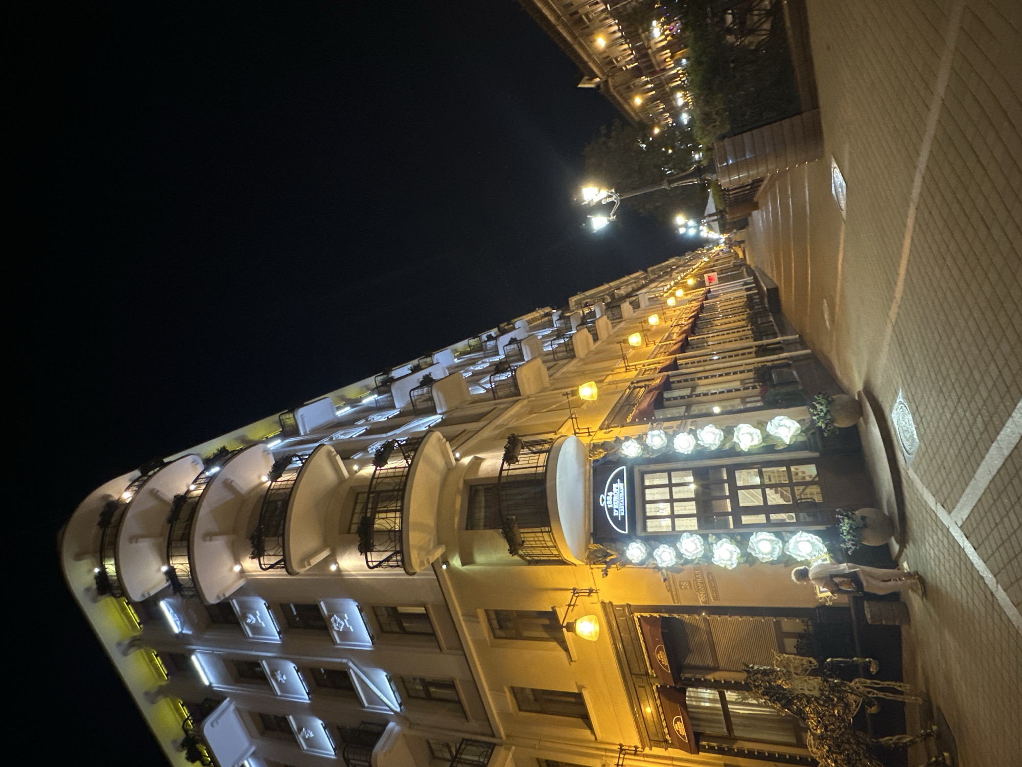 The image shows a street scene at night in Minsk, Belarus. A multi-story building with ornate balconies and illuminated windows lines the left side of the street. The building's facade is decorated with white floral-like lights around a large arched doorway. A woman in light-colored clothing is standing on the sidewalk in front of the building, looking at something in her hands. To her left is a large, decorative metal sculpture of a horse. The street is paved with cobblestones and illuminated by streetlights and the building's lights. The right side of the street shows another building with illuminated windows and awnings, and a line of trees and bushes. The sky is dark, indicating it is nighttime. The building on the left appears to be a restaurant or hotel, with signage visible that reads "1924 D'EUROPE RESTAURANT."