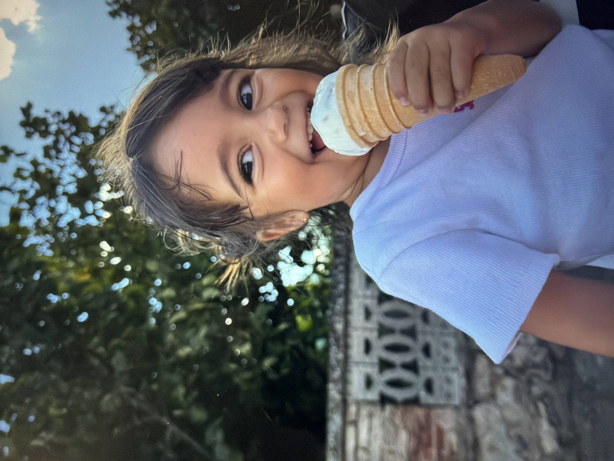 A young child with brown hair and eyes is holding a vanilla ice cream cone to their mouth. The child is wearing a light-colored shirt. The background includes foliage, a blue sky with clouds, and a brick wall with a decorative pattern. The location is Unknown, Unknown.