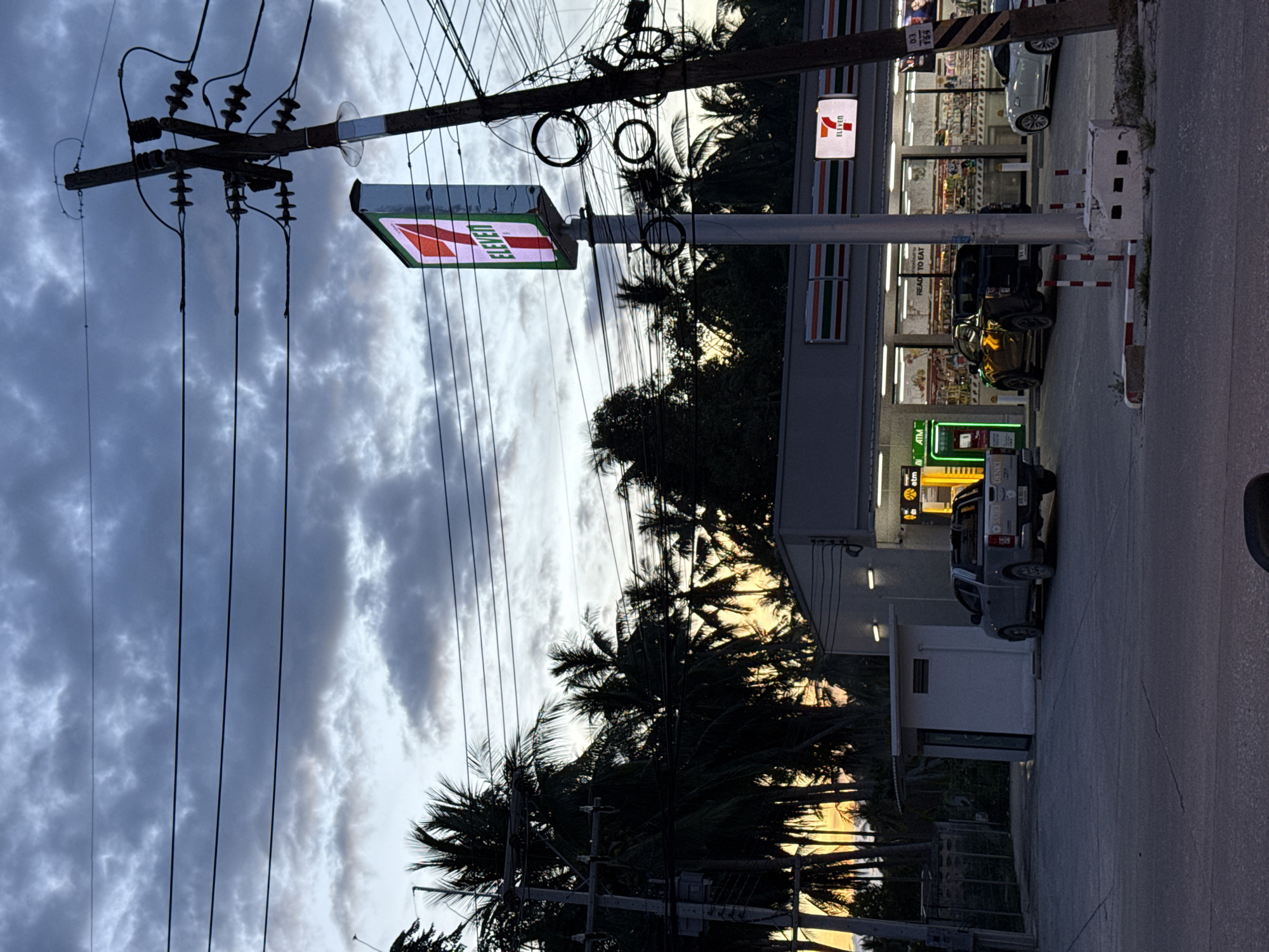 The image depicts a 7-Eleven convenience store at dusk in Baan Nai Suan, Thailand. In the foreground, power lines are strung across a utility pole. Two 7-Eleven signs are visible, one perpendicular to the road and another attached to the store building. Palm trees obscure some of the background. Several vehicles are parked near the storefront.