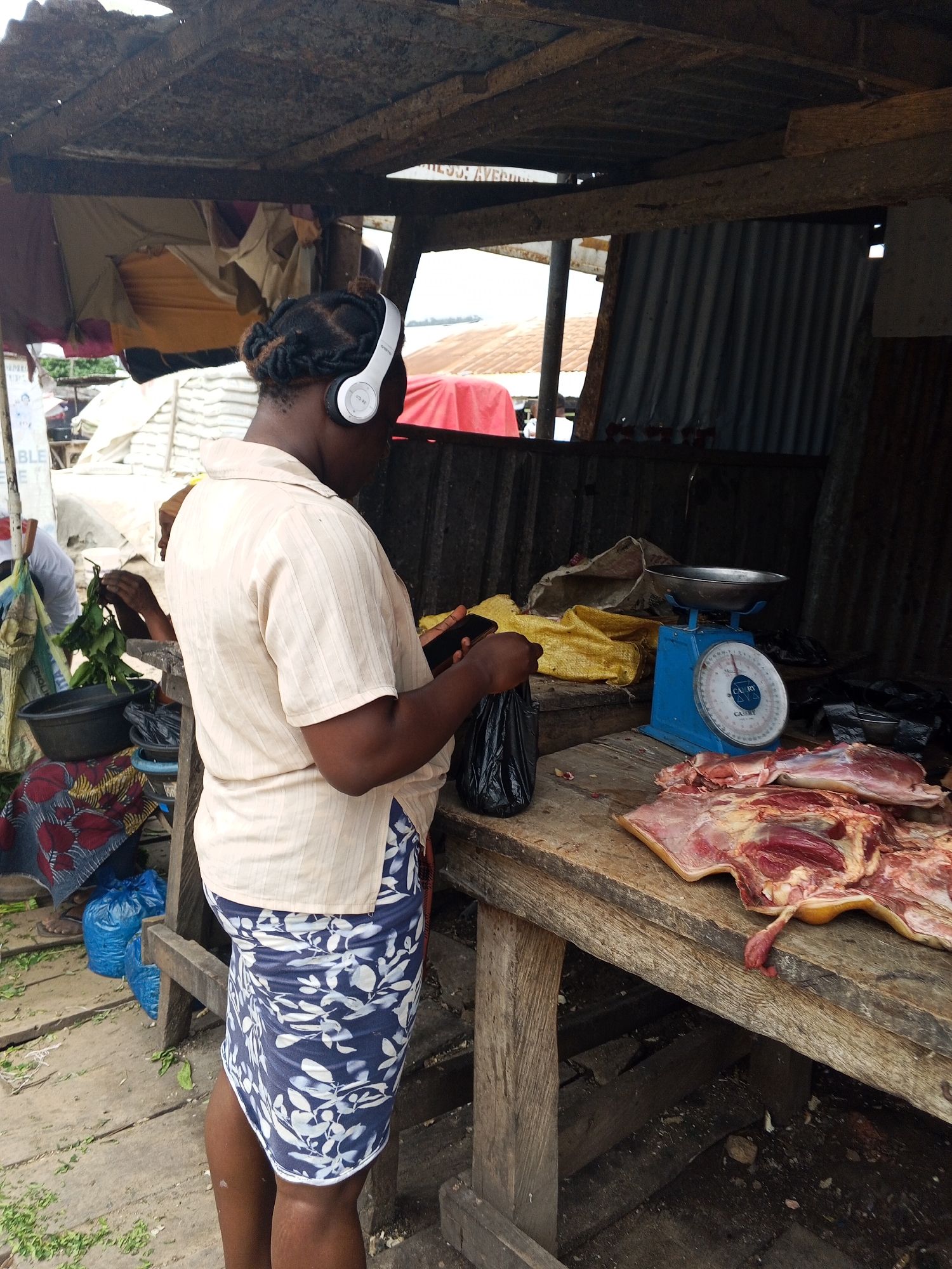 A medium shot shows a person with braided hair wearing white headphones and a light-colored, collared shirt with vertical stripes, and a blue and white floral skirt, standing at a wooden market stall. The person is holding a black plastic bag in their left hand and a smartphone in their right hand. In front of them on the wooden table is a blue mechanical scale with a silver pan, and a large piece of raw meat with visible fat and muscle. To the left of the scale, there are yellow and grey cloths. Behind the person, the stall structure is visible with a corrugated metal roof and wooden beams. Other market stalls and items are faintly visible in the background. The ground is made of wooden planks with some scattered green debris.