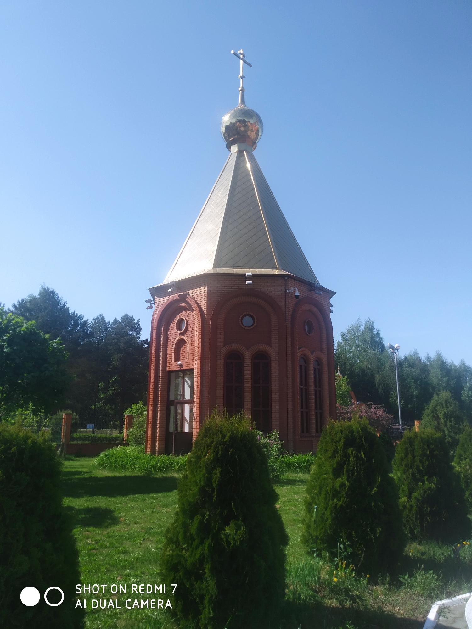 The image depicts a small, single-story brick Orthodox church or chapel with a hexagonal base. The roof is conical, covered in gold-colored metal tiles and surmounted by a golden onion dome with a cross on top. The building features arched windows and decorative brickwork. The structure is surrounded by a well-maintained lawn and several small evergreen trees. The background consists of a line of larger trees, possibly marking the edge of a forest or park. The sky is clear and blue. The image was taken in Chaikovsky, Russia.
