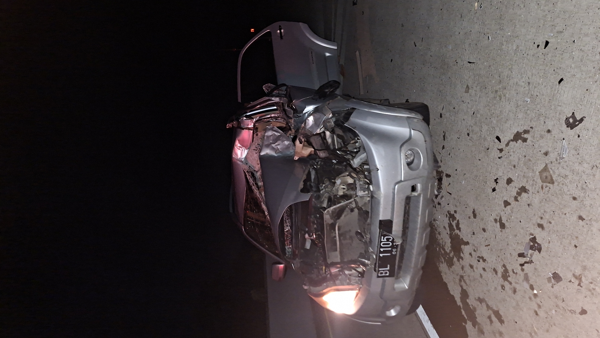 A silver car has been in a severe accident, with its front end completely crushed. The car is lying on its side on a concrete surface, possibly a road or a roadside. Debris from the accident is scattered around the vehicle. The car's hood is open, revealing further damage to the engine compartment. A license plate is visible on the front of the car, reading "BL 1105" and "06-27". The scene appears to be illuminated by the car's headlights and possibly by artificial light sources, suggesting it is nighttime or very dark. The background is dark, indicating it is likely night. The concrete surface is stained with what appears to be liquids and small fragments, typical of an accident scene. The overall impression is one of destruction and chaos resulting from a significant collision. The location is Baksukon, Indonesia.